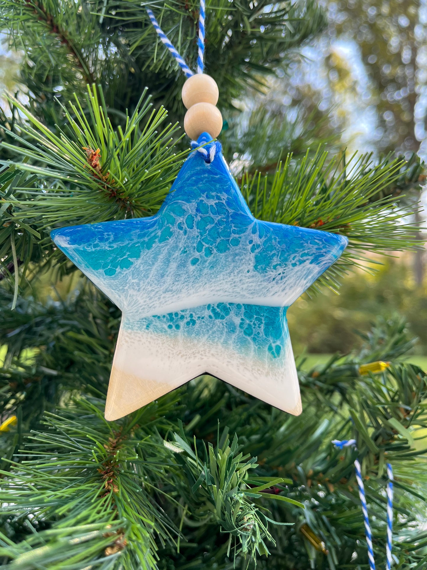 Star-shaped wooden Christmas ornament covered in blue and teal ocean resin art resembling the waves of the ocean.  Finished with blue and white baker's twine and two wooden beads