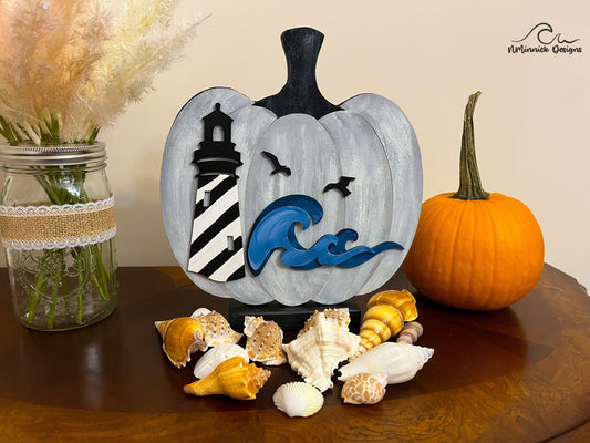 a white and gray wooden standing pumpkin with a lighthouse scene on the front.  The lighthouse is white with black swirls resembling Hatteras Lighthouse.  There are 3D waves in dark and light blue and two black seagulls flying over the ocean.  Coastal pumpkin is sitting on a table next to seashells, a real pumpkin, and a flower arrangement.