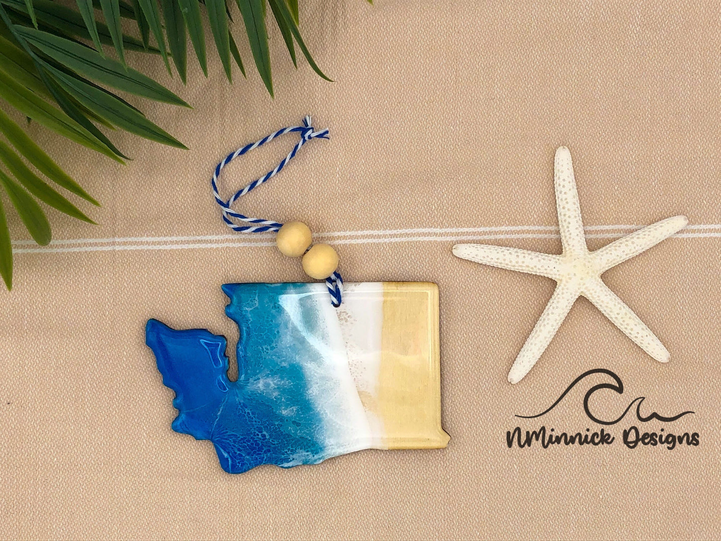 1/8 inch basswood ornament in the shape of Washington State. Covered in multiple layers of blue, teal, and white epoxy resin to create a one of a kind ocean beach scene. Finished with a bakers twine hanger and two wooden beads.