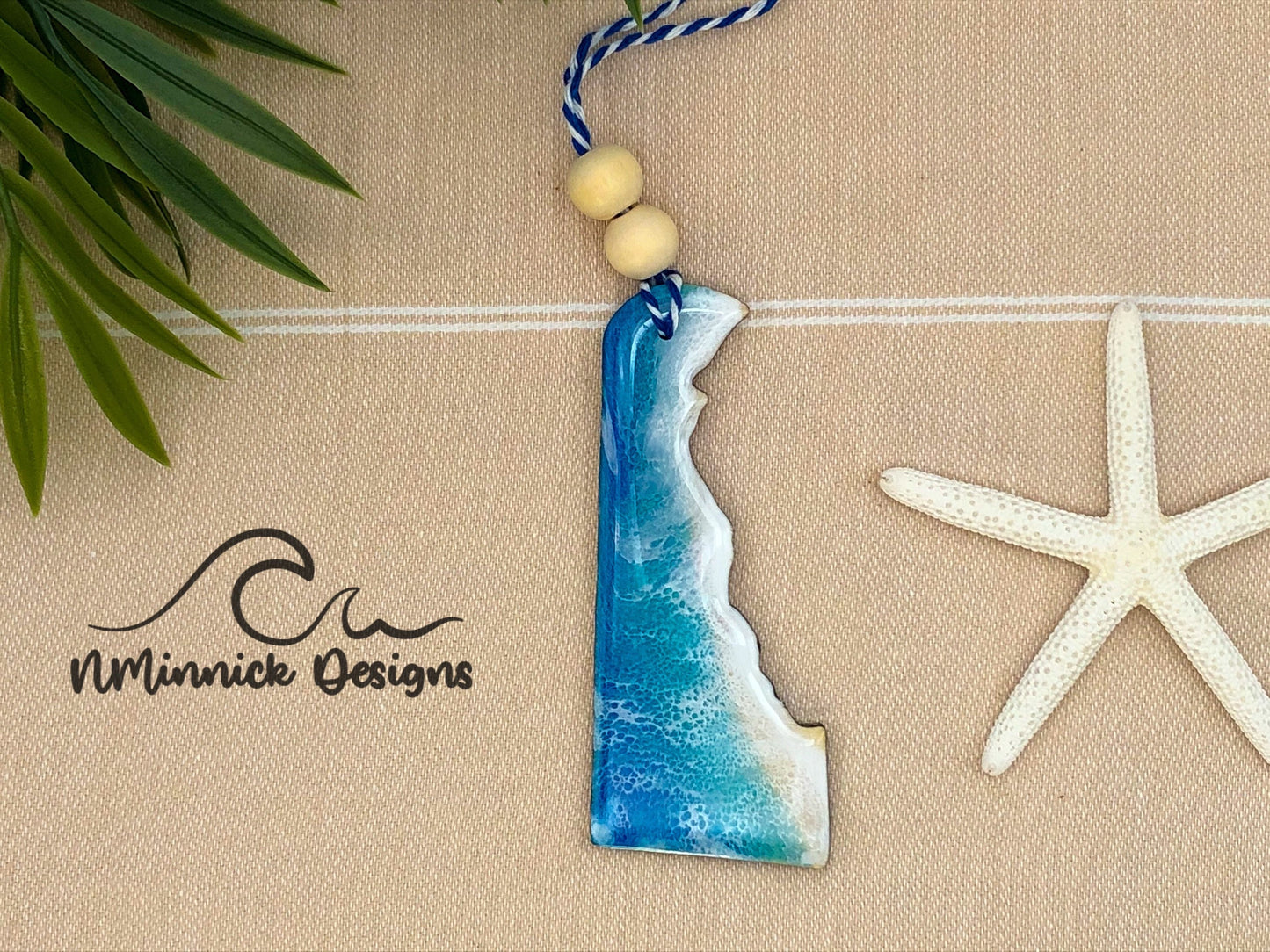 Delaware shaped wood ornament covered in blue, teal, and white resin to create a one of a kind beach scene. Approx. 4 inches long and finished with bakers twine and two wooden beads.