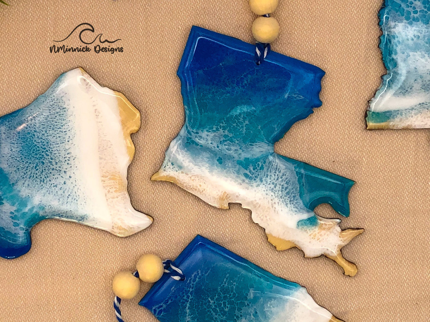 4 to 4.5 inch laser cut ornament in shape of Louisiana, coated in blue, teal and white resin to create a one of a kind beach scene. Finished with bakers twine and two wooden beads.