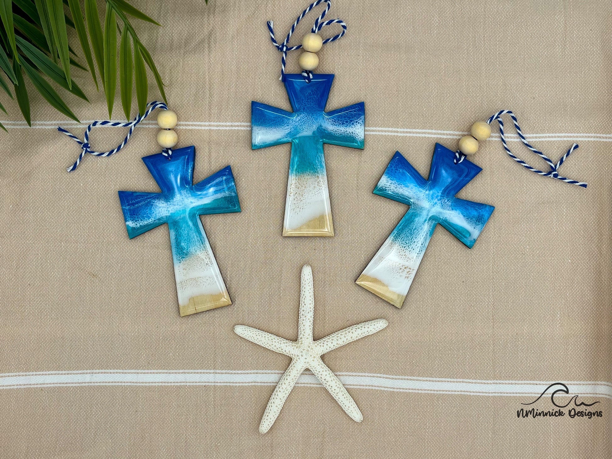 Three cross shaped wooden Christmas ornaments covered in blue ocean resin art, resembling ocean waves. Finished with baker&#39;s twine and two wooden beads. Laying on a tan beach towel next to a natural starfish and palm leaves.