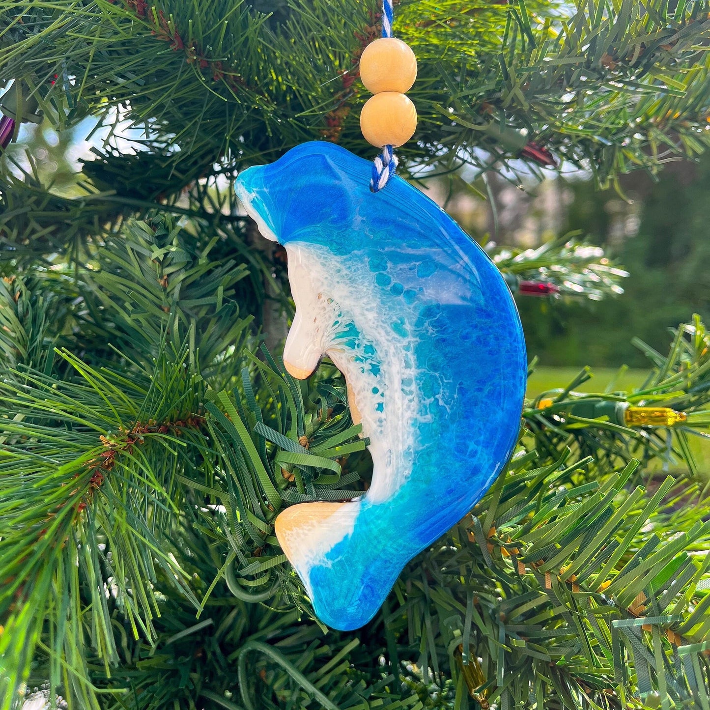A manatee shaped wooden Christmas ornament covered in blue ocean resin art, resembling the waves of the ocean. Finished with baker&#39;s twine and two wooden beads and hanging on a Christmas tree.