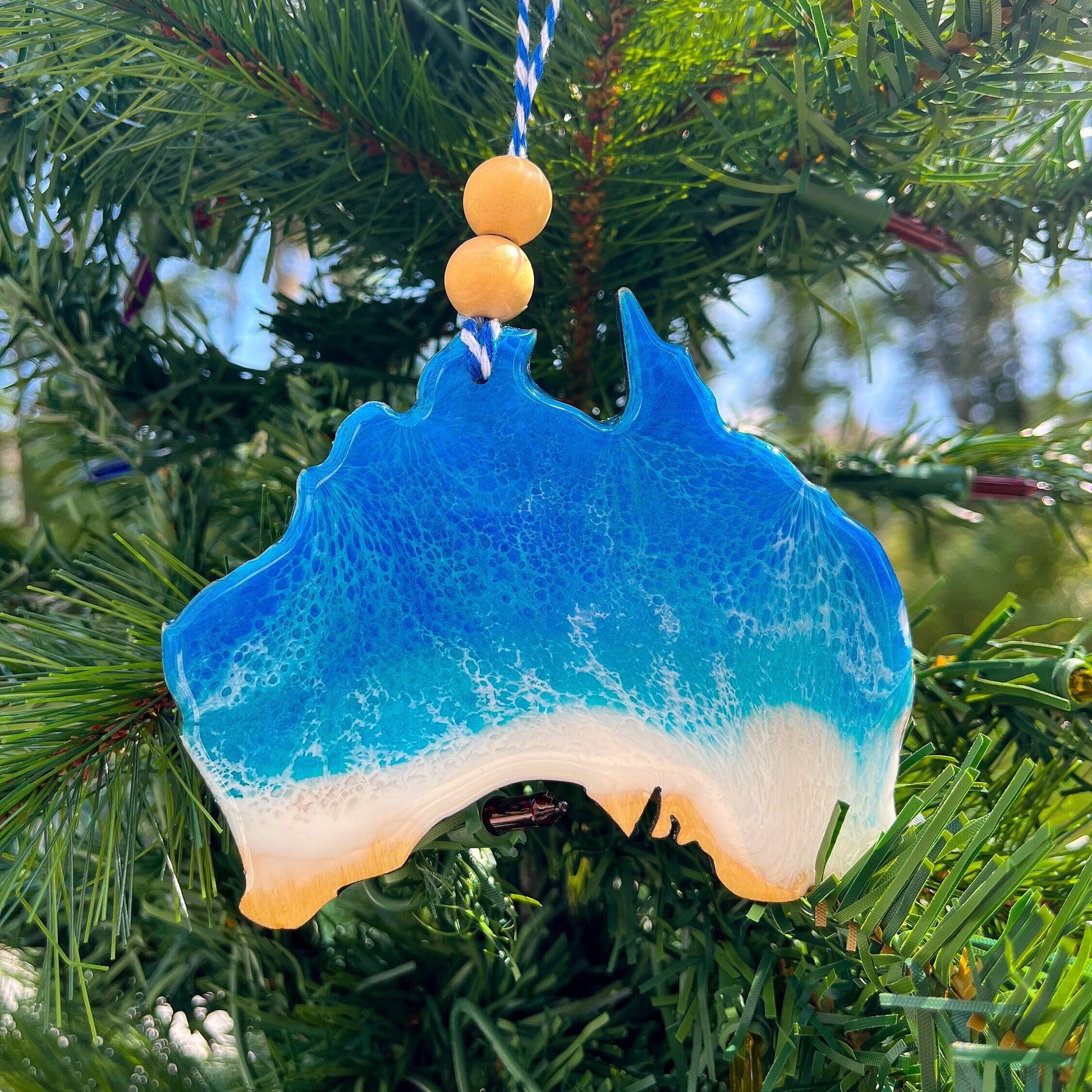 An Australia shaped wooden Christmas ornament covered in blue ocean resin art, resembling the waves of the ocean. Finished with baker&#39;s twine and two wooden beads and hanging on a Christmas tree.