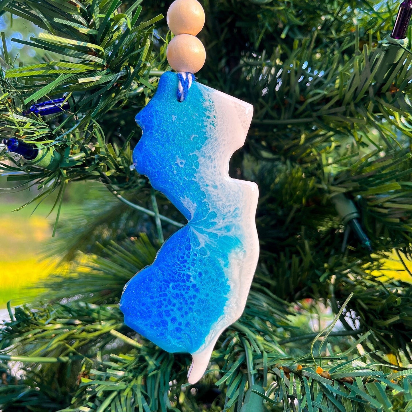 New Jersey-shaped wooden Christmas ornament covered in blue and teal colored ocean resin art resembling the waves along New Jersey&#39;s Atlantic Coast. Finished with blue and white baker&#39;s twine and two wooden beads.