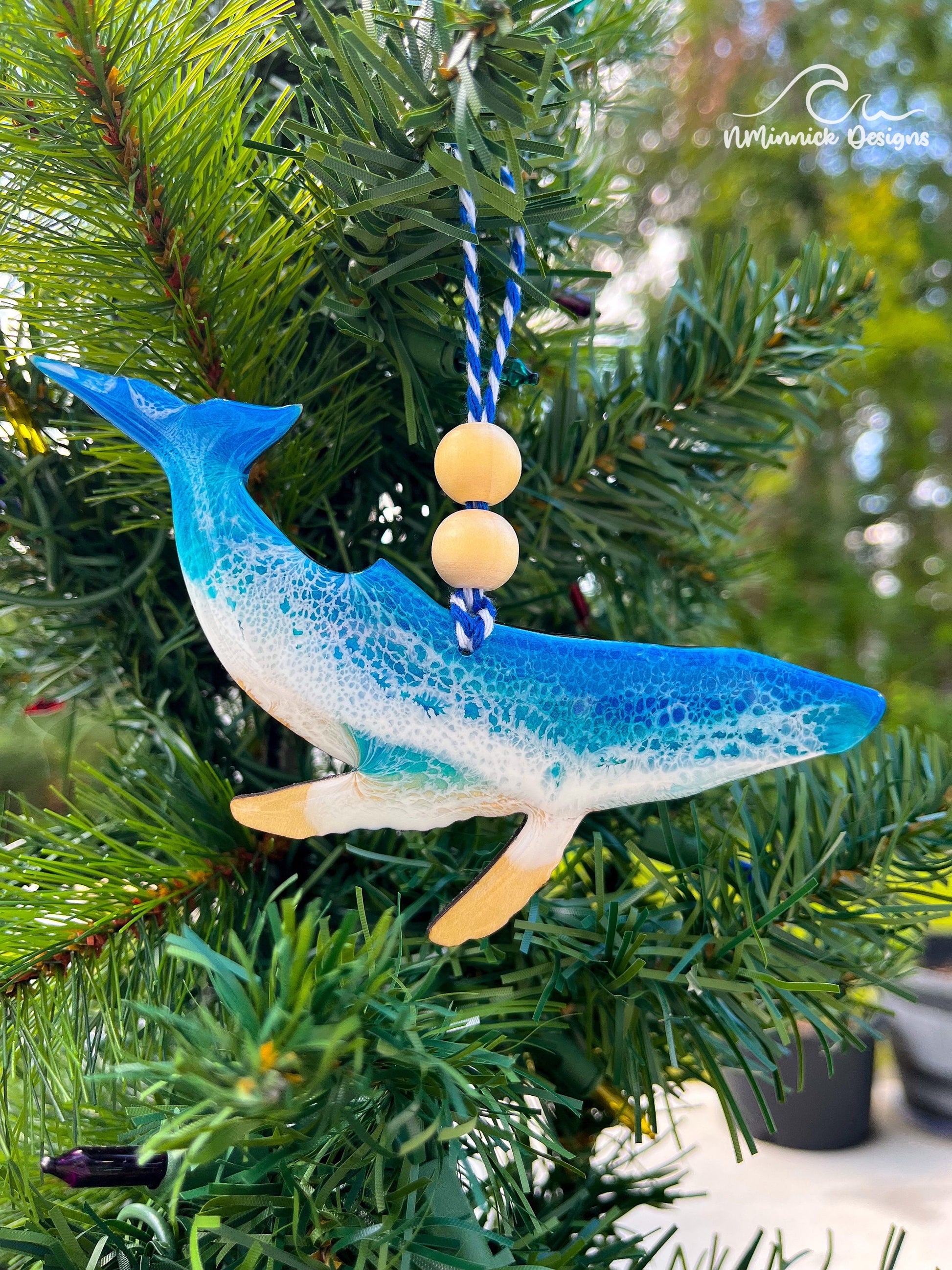 a humpback whale shaped wooden Christmas ornament covered in ocean blue and Okinawa blue ocean resin art resembling ocean waves. Finished with baker&#39;s twine and two wooden beads and hanging on a Christmas tree.
