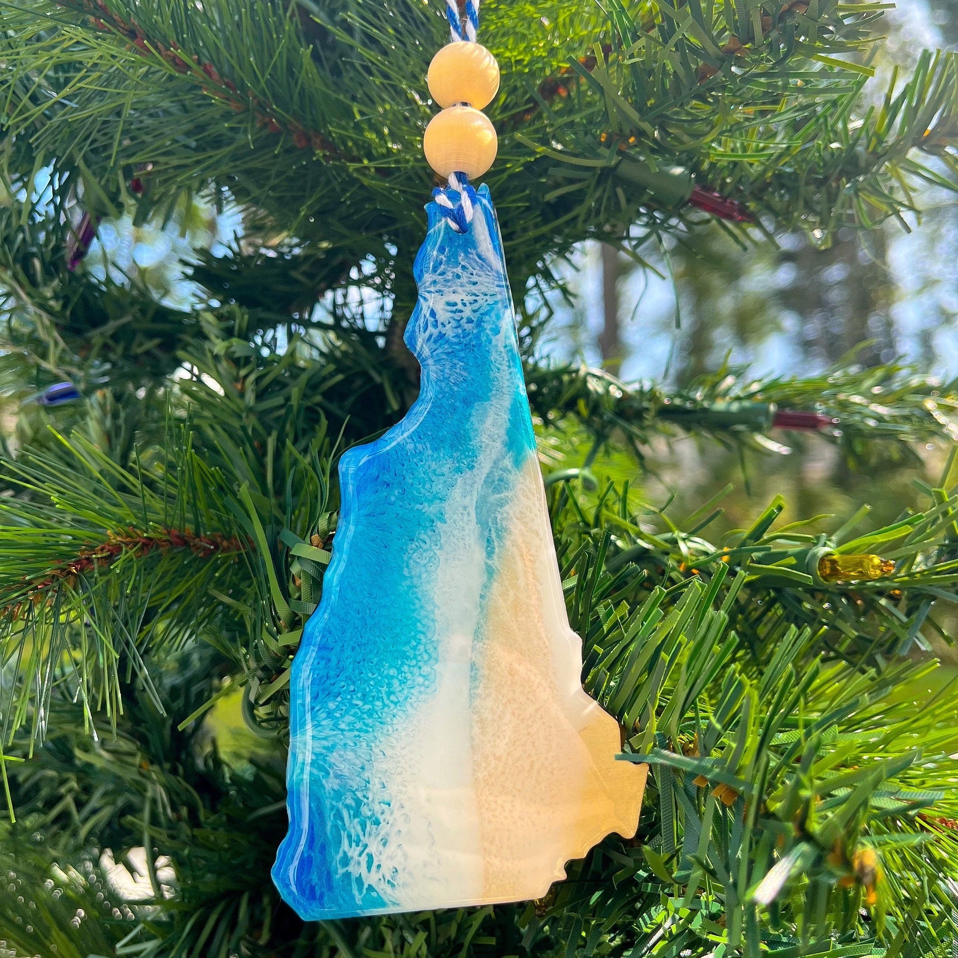 a new hampshire state shaped wooden Christmas ornament covered in ocean blue and okinawa blue ocean resin art resembling ocean waves against the shore. Finished with baker&#39;s twine and two wooden beads and hanging on a christmas tree.