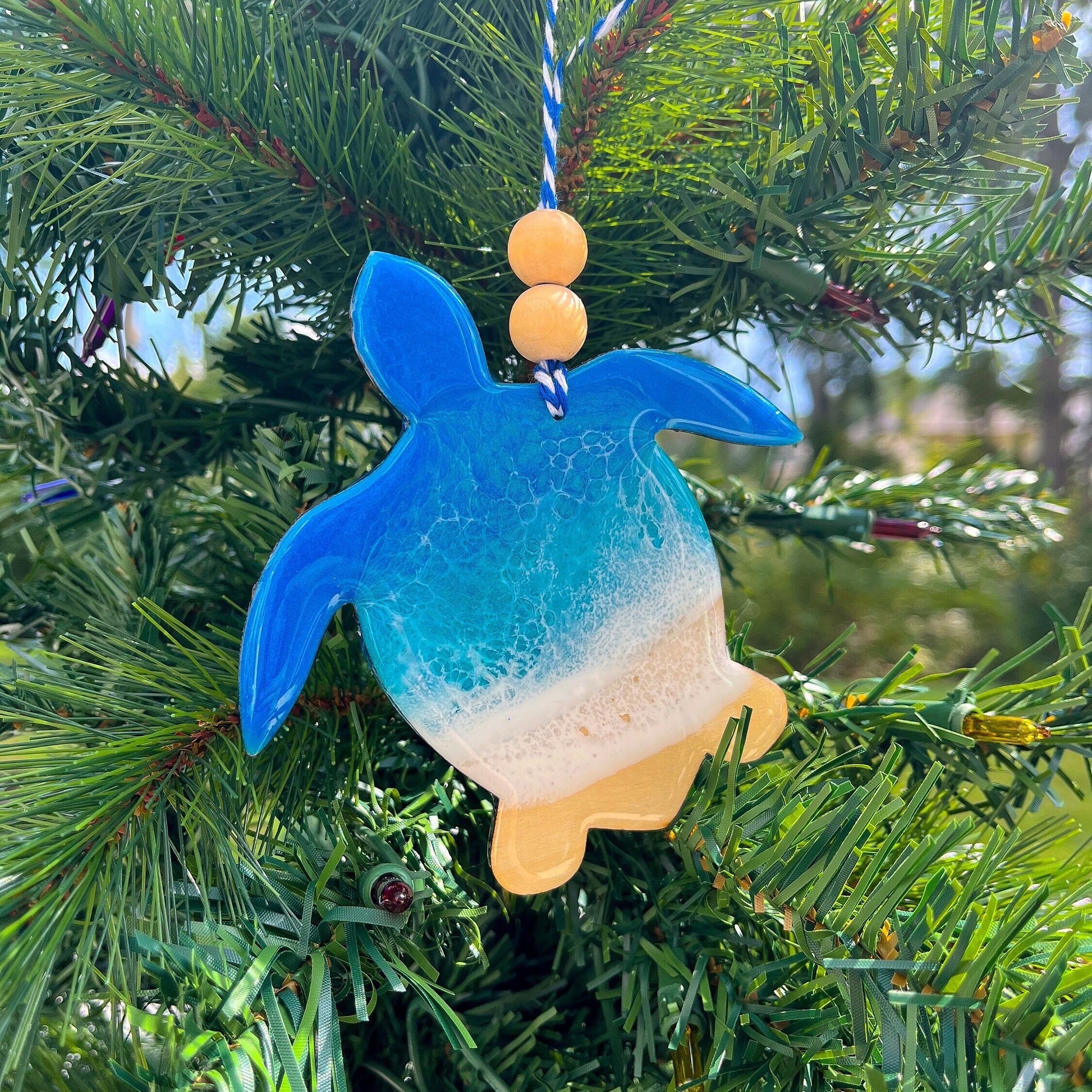 a Sea Turtle shaped wooden Christmas ornament covered in blue ocean resin art, resembling the waves of the ocean. Finished with baker&#39;s twine and two wooden beads and hanging on a Christmas tree.