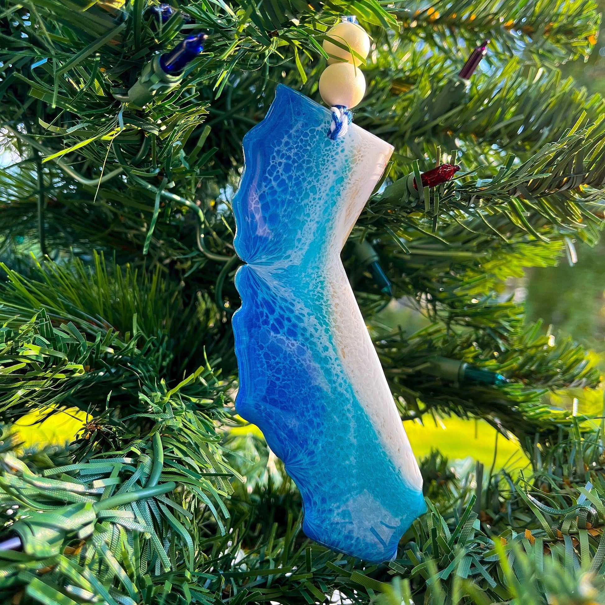 A california shaped wooden christmas ornament covered in layers of blue and teal ocean resin art resembling the waves of the ocean. Finished with baker&#39;s twine and two wooden beads and hanging on a christmas tree.