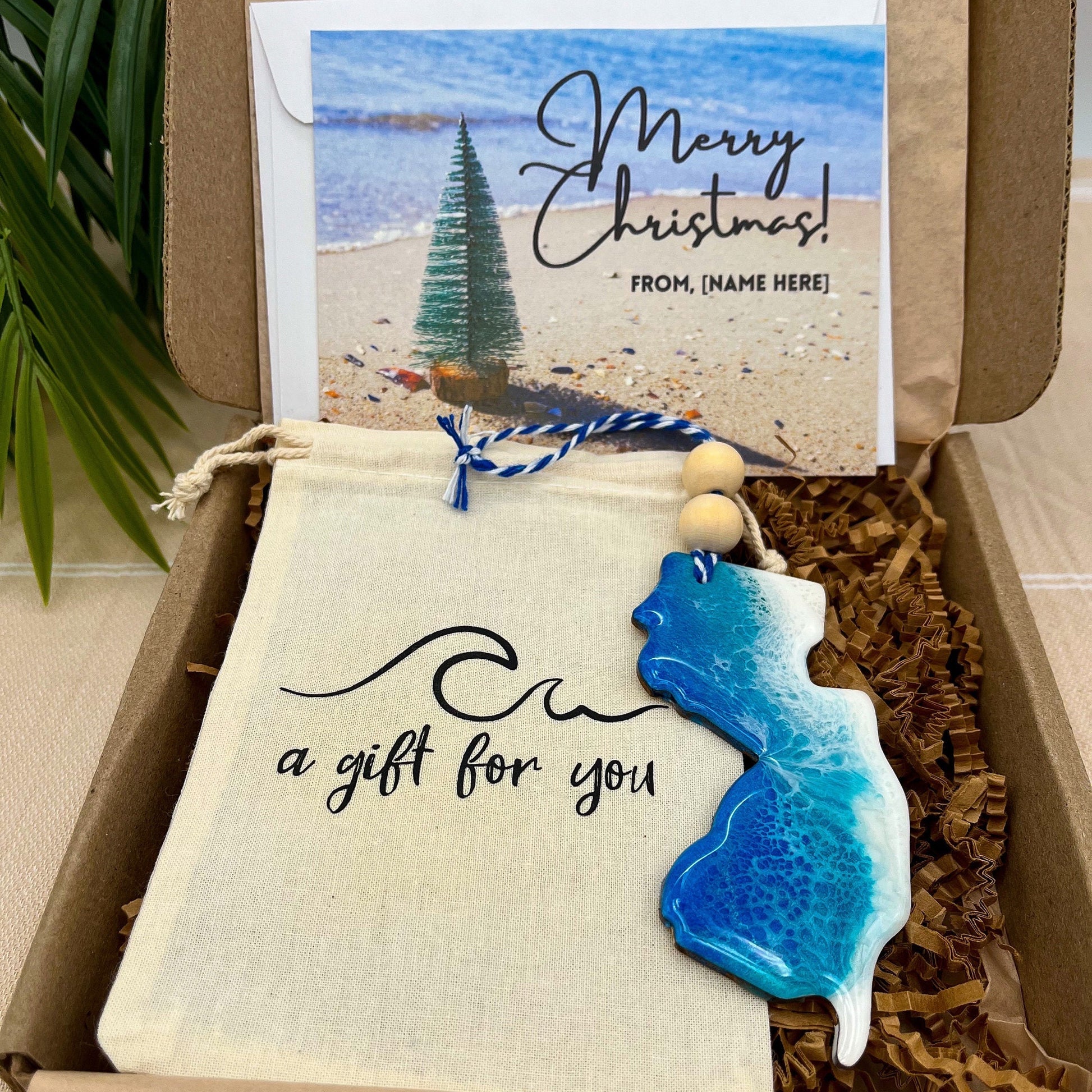 A New Jersey shaped wooden christmas ornament with ocean resin art, resembling the waves of the ocean. In a box with an ornament keepsake bag with a wave that says &quot;a gift for you&quot; and a Merry Christmas custom card.
