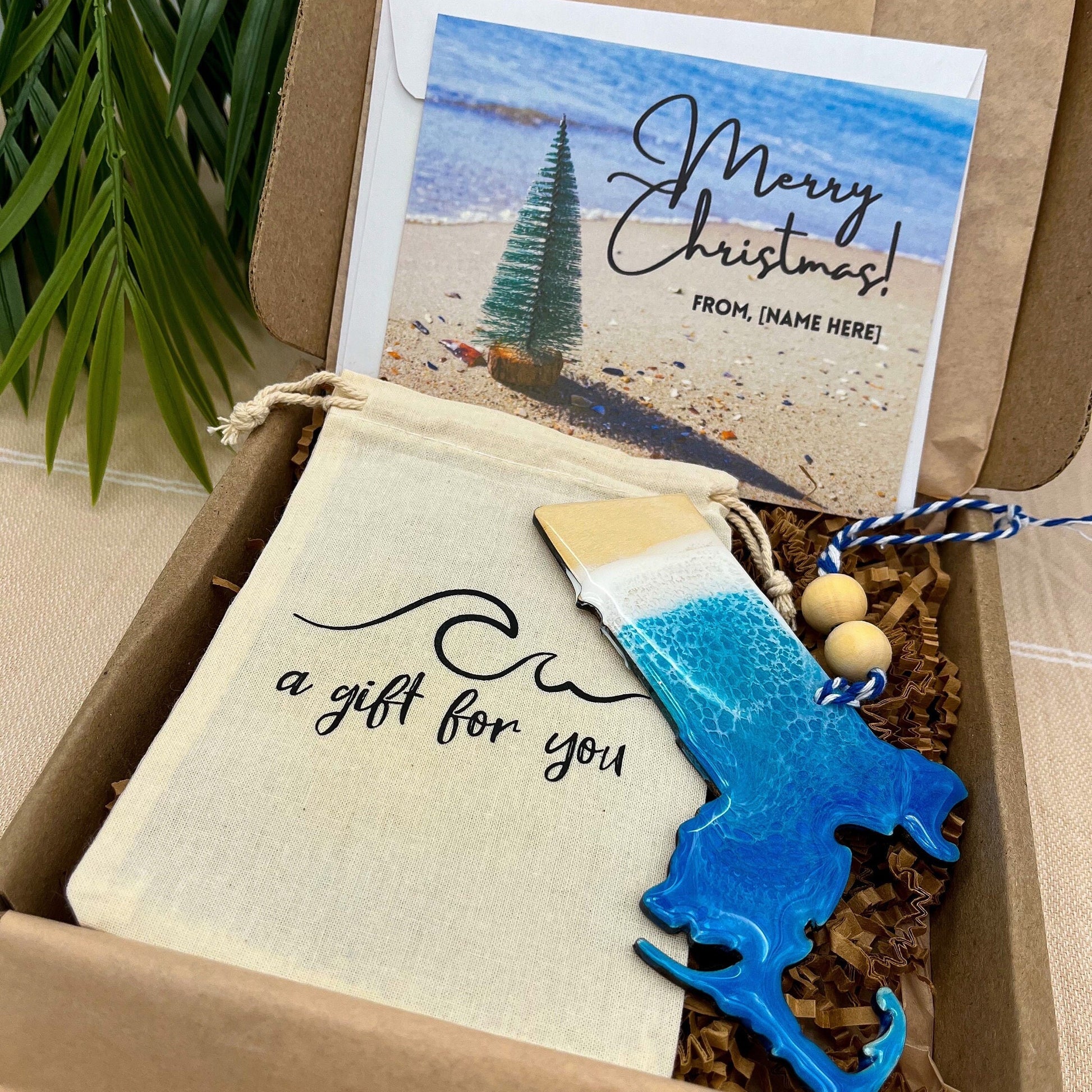 A Massachusetts shaped wooden christmas ornament with ocean resin art, resembling the waves of the ocean. In a box with an ornament keepsake bag with a wave that says &quot;a gift for you&quot; and a Merry Christmas custom card.