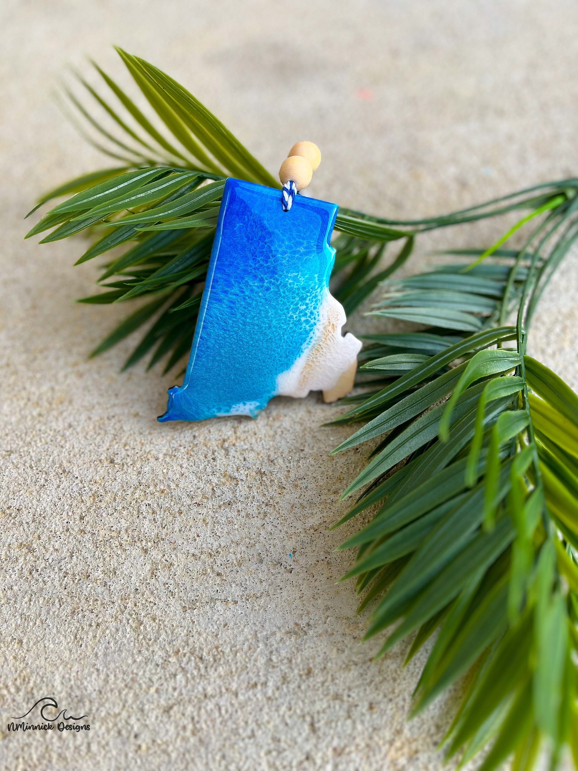 Rhode Island shaped ornament with ocean resin art laying against palm leaves.