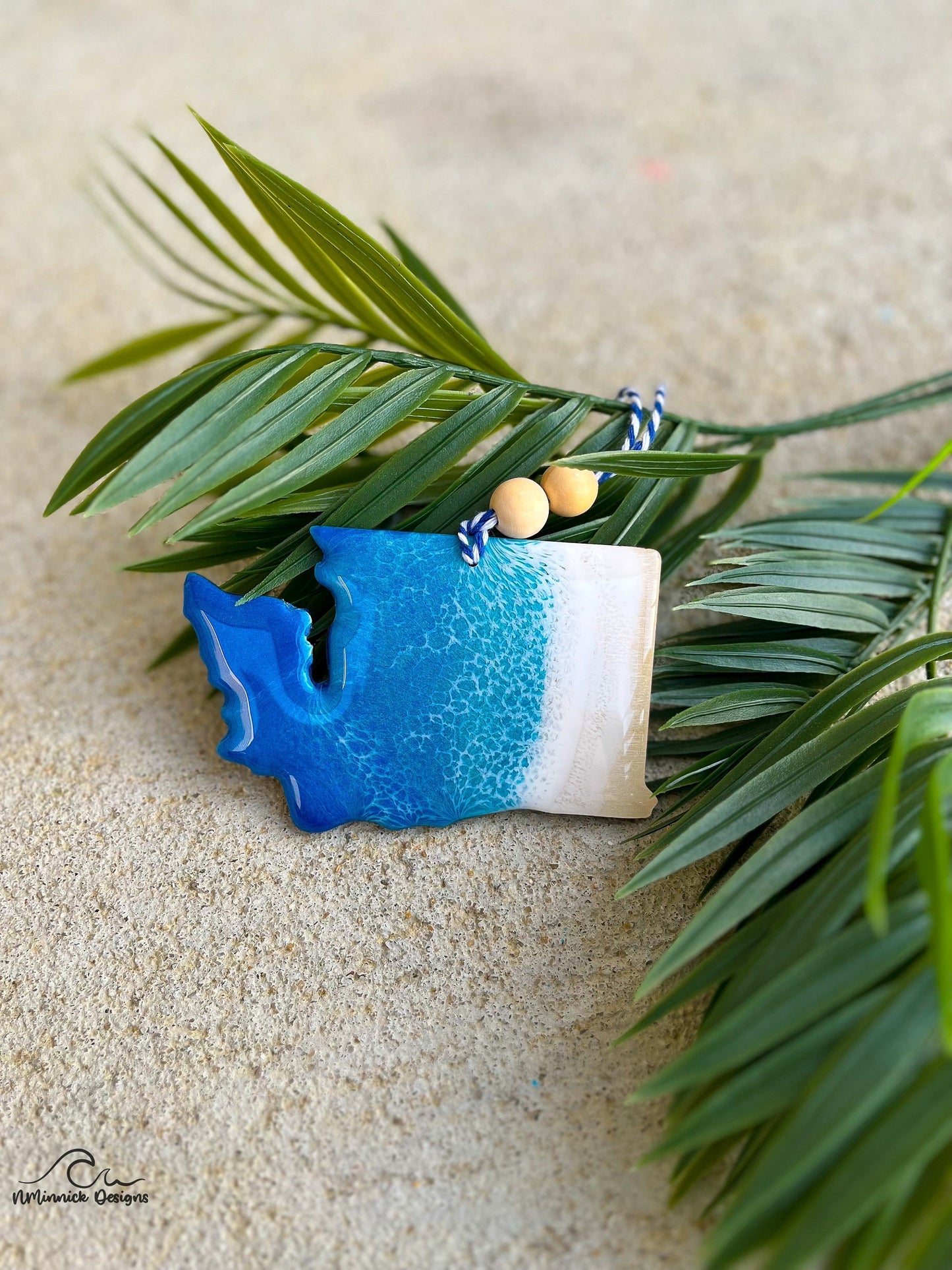 Washington state shaped ornament with ocean resin art laying against palm leaves.