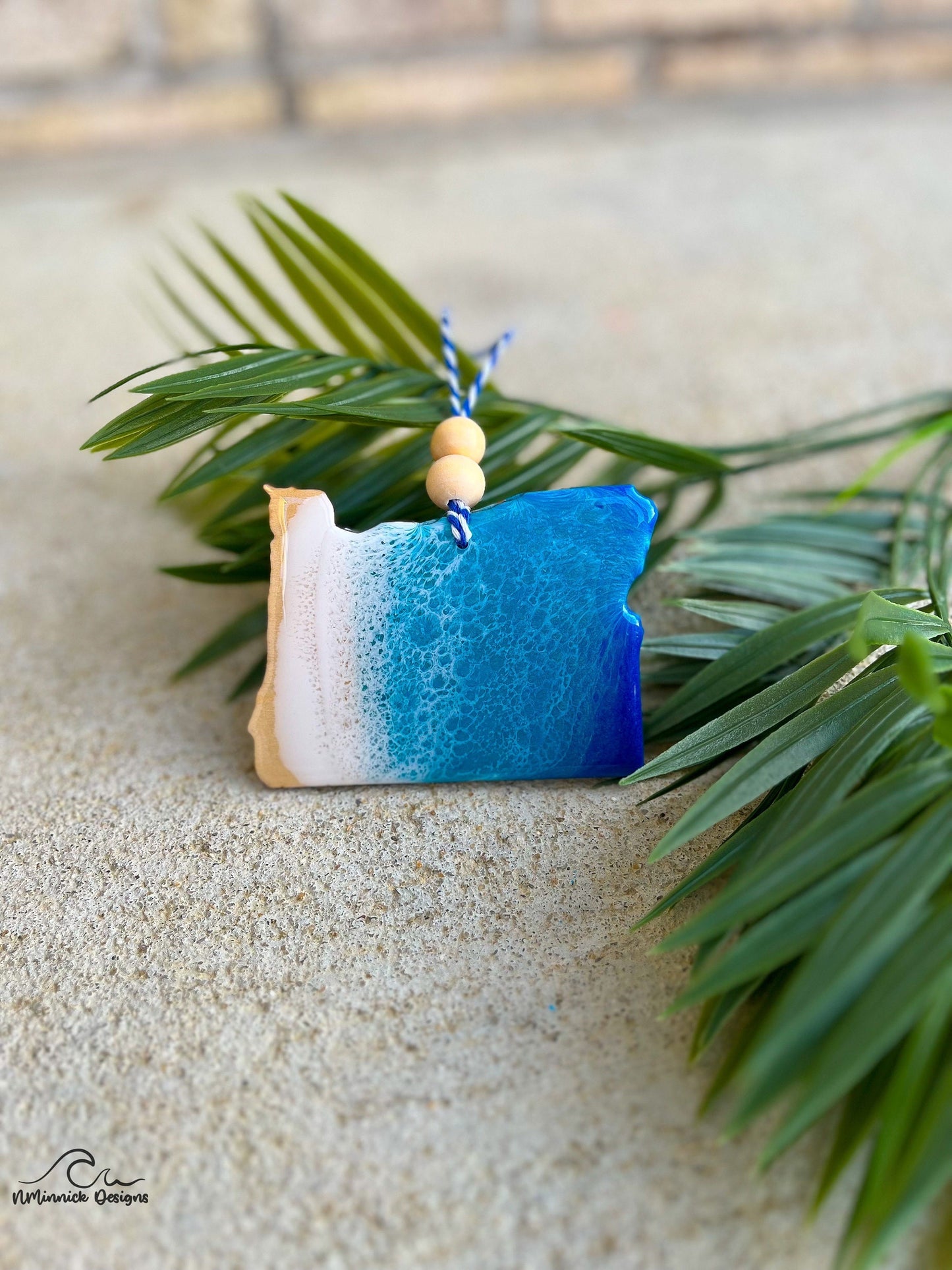 Oregon shaped wooden ornament with ocean resin art laying against palm leaves.
