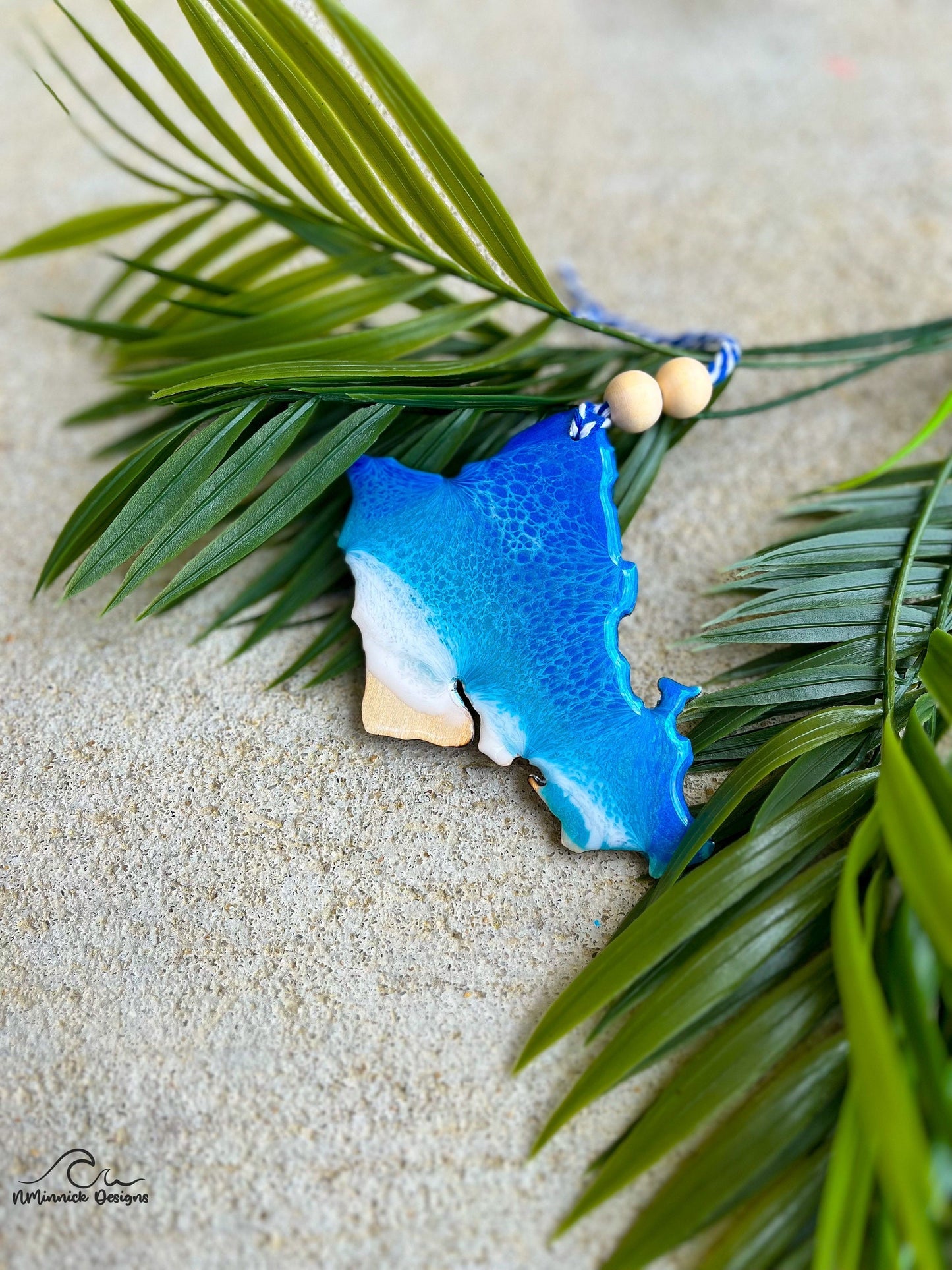 An Oahu shaped ornament with ocean resin art laying against palm leaves.