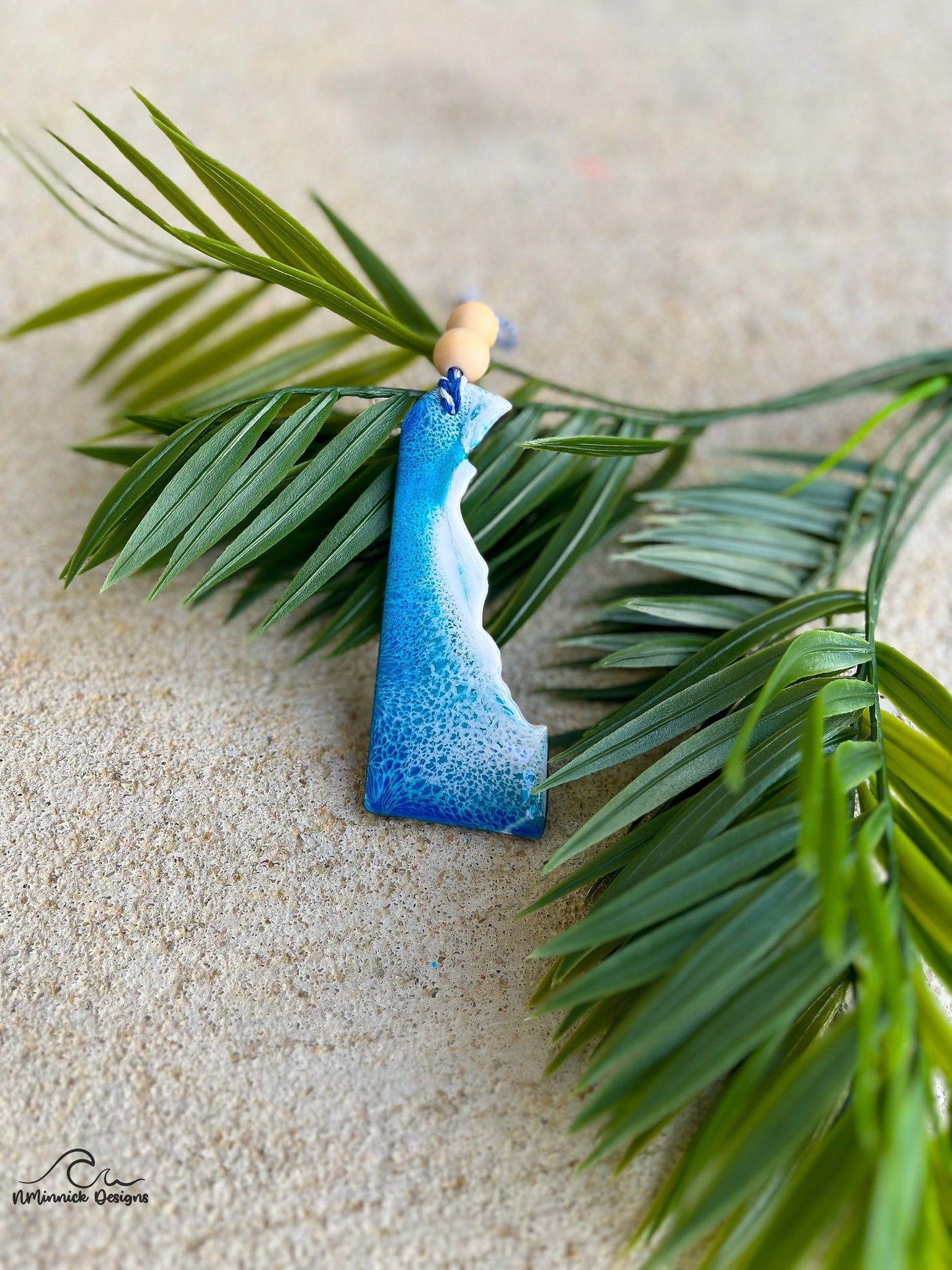 A delaware shaped wooden ornament with blue and white ocean resin art, finished with a strand of baker&#39;s twine and two wooden beads.