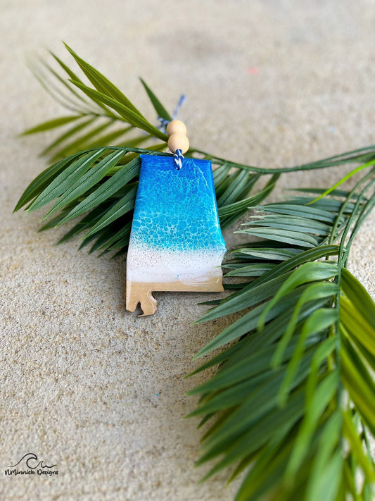 Alabama ornament with ocean resin art laying against palm leaves.