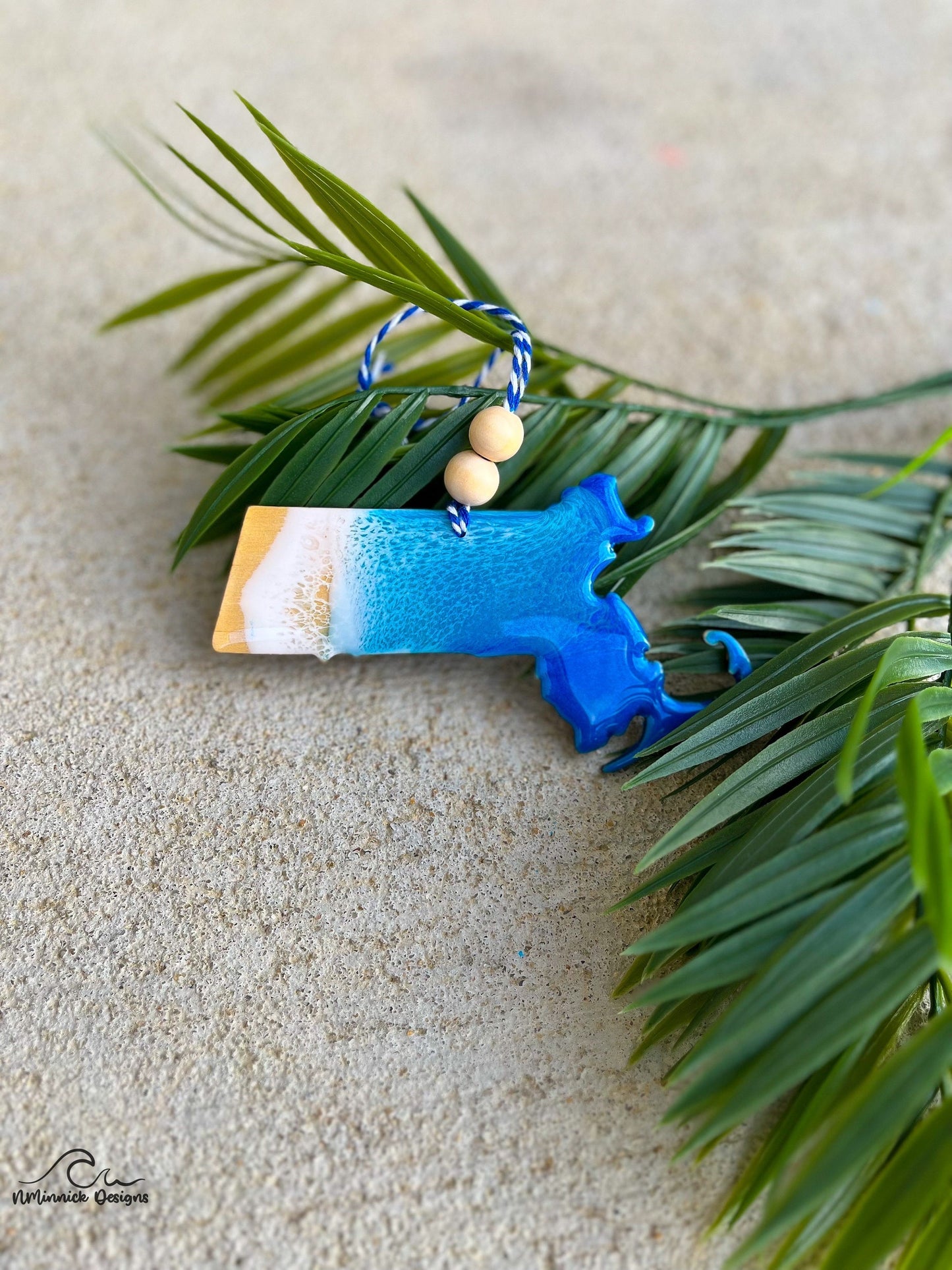 Massachusetts Ornament with ocean resin art laying against palm leaves.