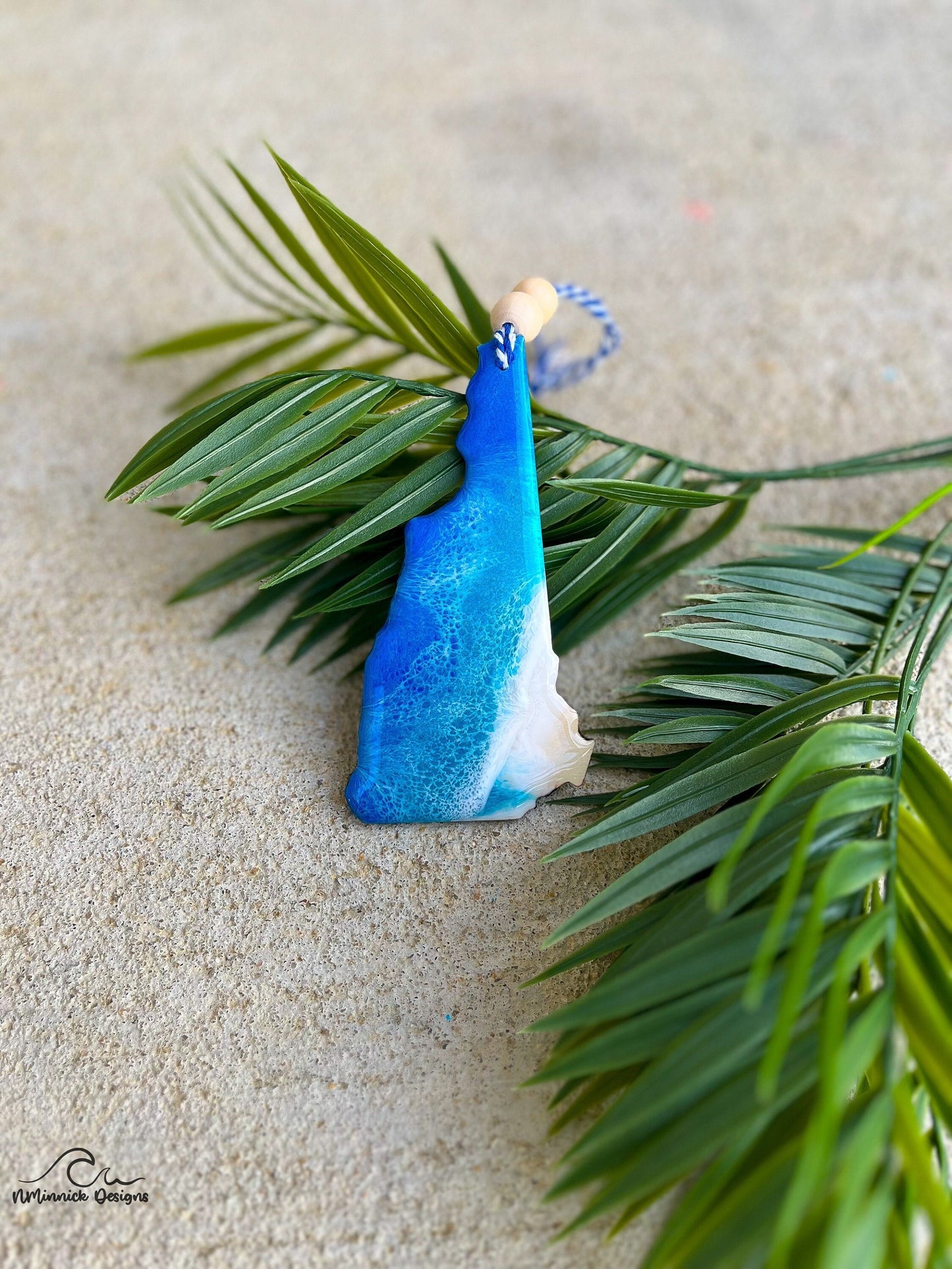 New Hampshire Ornament with ocean resin art laying against palm leaves.