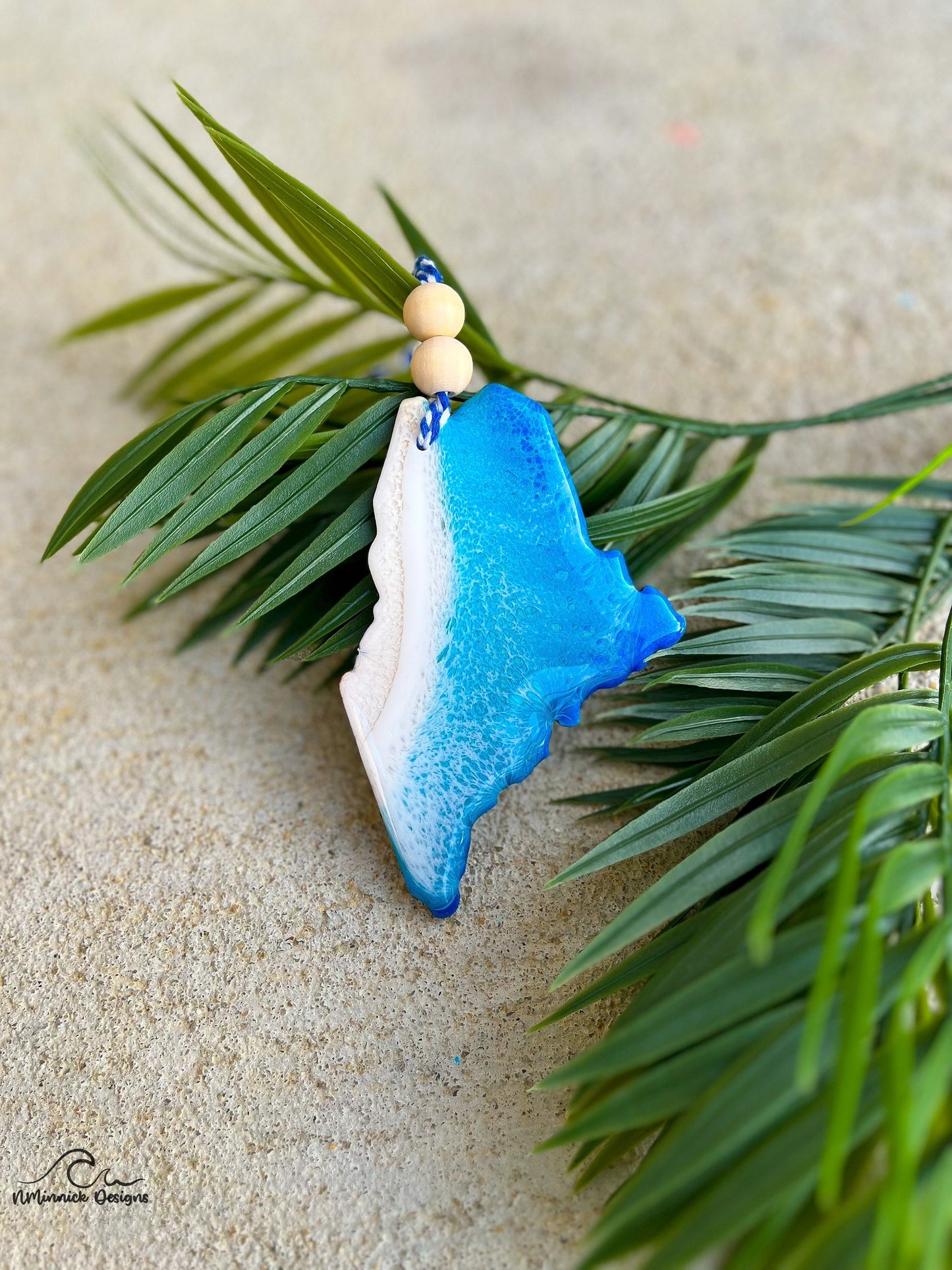 Maine shaped wood and resin ornament made to look like ocean waves. Laying against palm leaves.