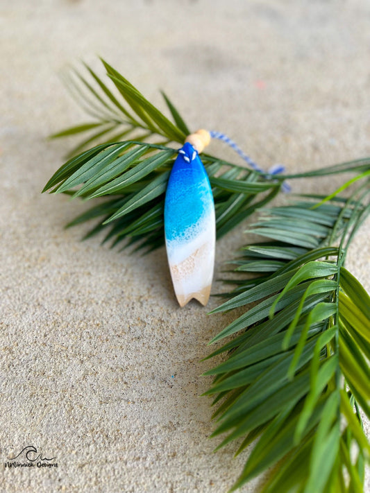 Surfboard ornament with ocean resin art laying against palm leaves.