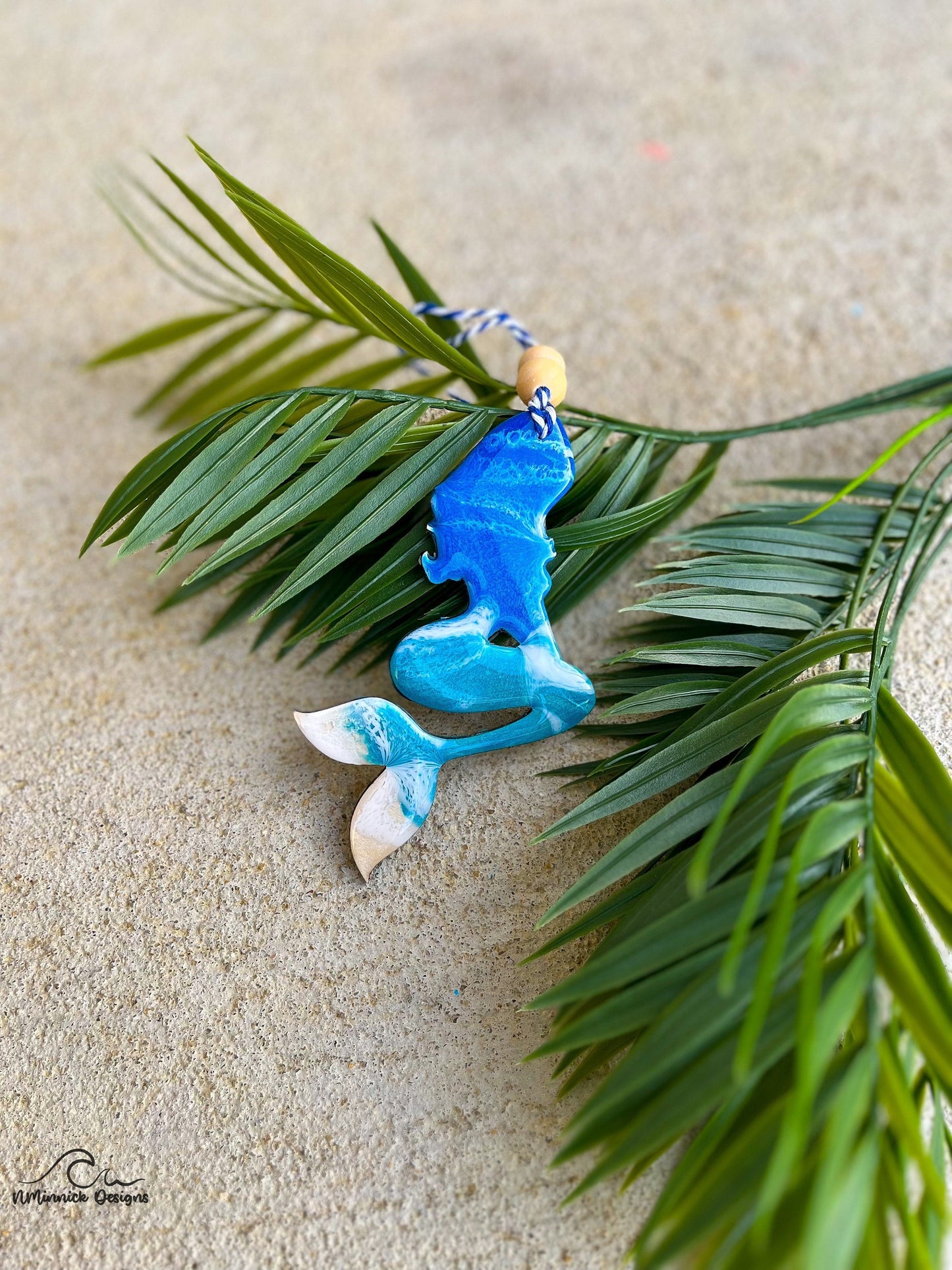 Mermaid shaped wood and resin ornament with ocean waves and laying against palm leaves.