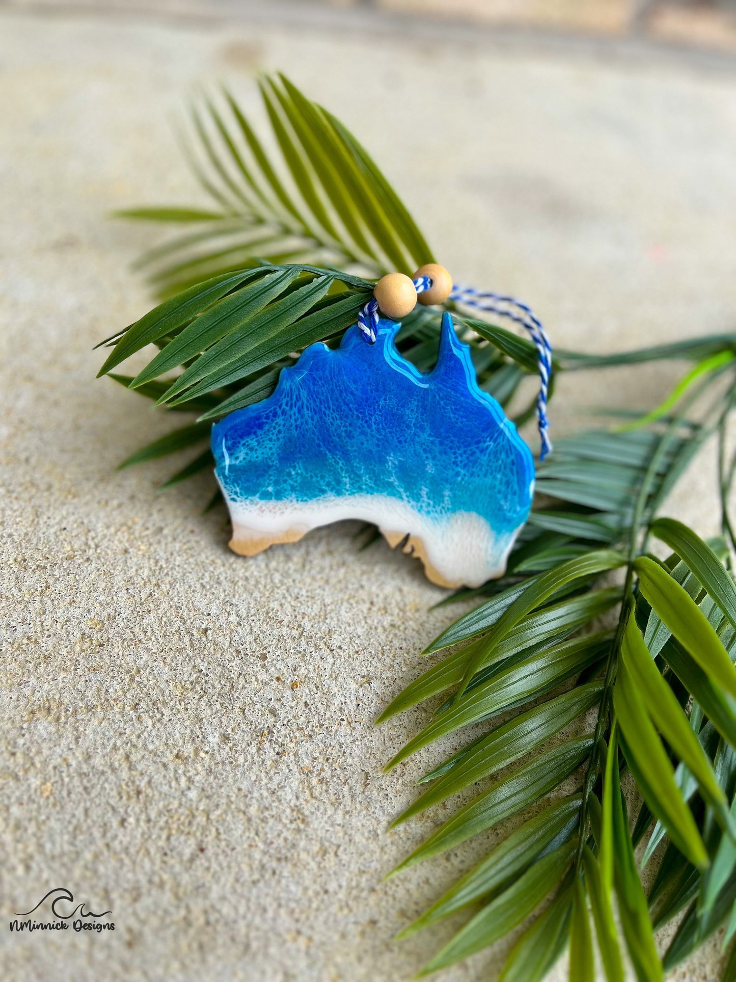 Australia shaped wood and resin ornament with ocean waves and laying against palm leaves.
