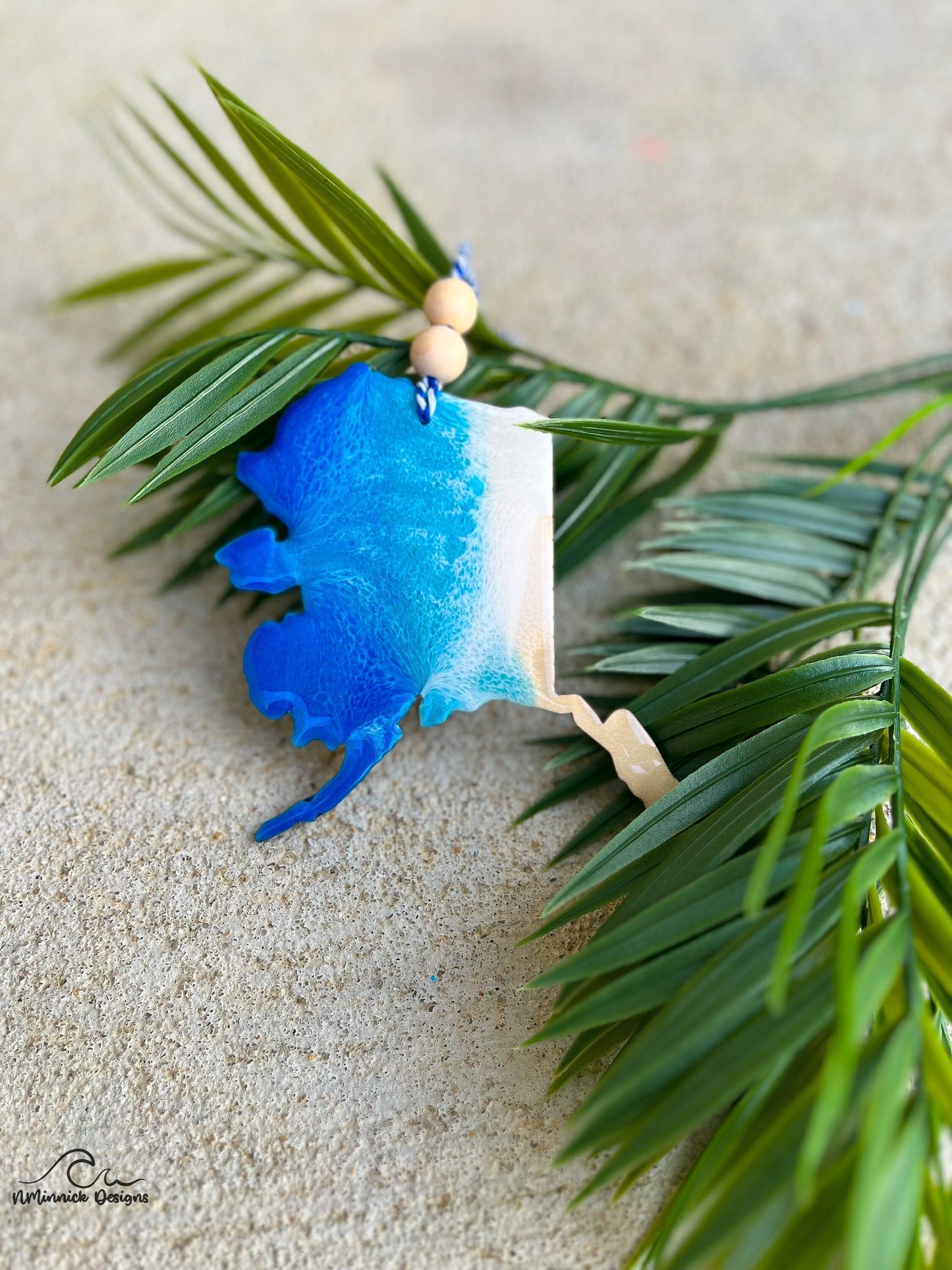 Alaska shaped wood and resin ornament with ocean waves laying against palm leaves.