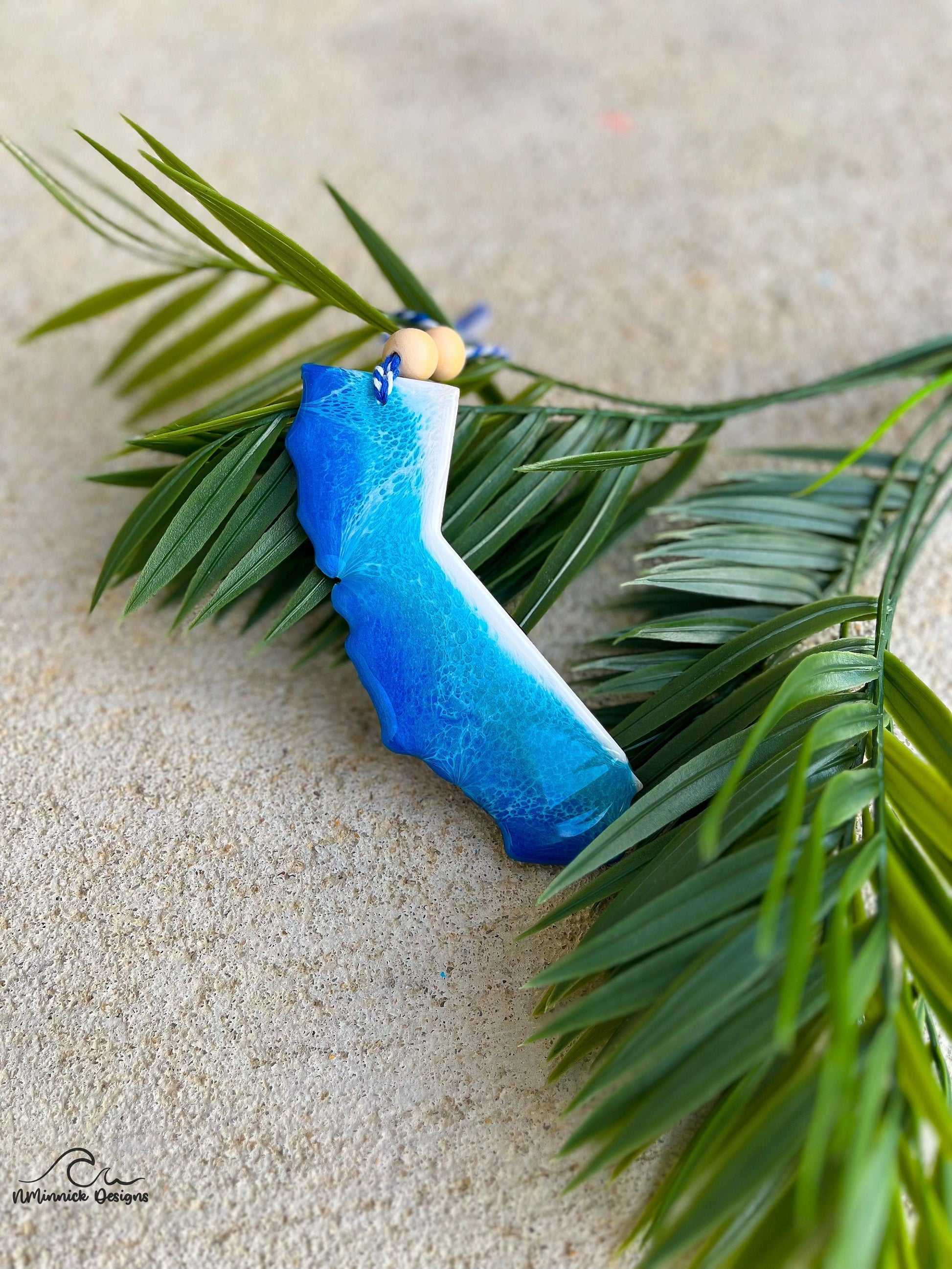 California shaped ornament with ocean wave art laying against palm leaves.