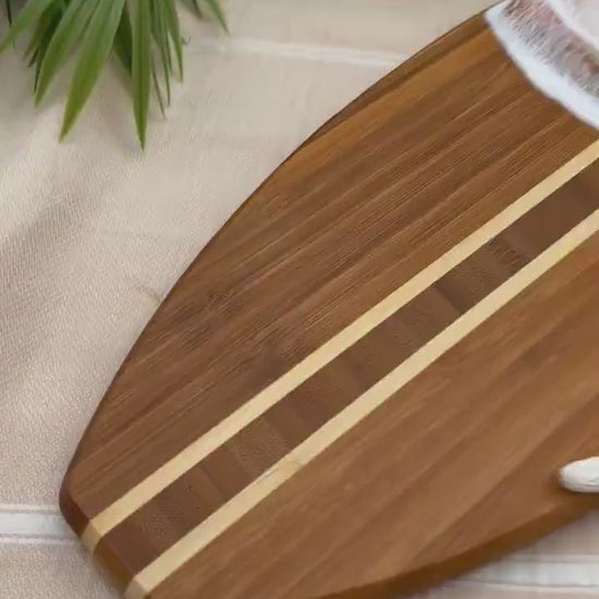 Surfboard-shaped bamboo cutting board with the top third of the board covered in a coral pink and gold tinted ocean resin art resembling the waves of the ocean. Laying on a tan beach towel next to a natural starfish (not included).