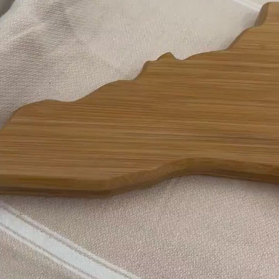 North Carolina-shaped charcuterie board with dark blue and teal ocean resin art along the atlantic coast of the state, resembling the waves of the ocean. Sitting up against a wooden walkway with the Bodie Island Lighthouse in the background.