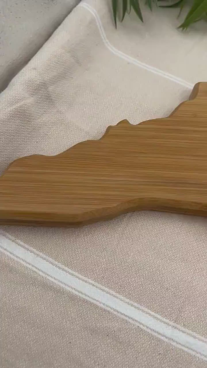North Carolina-shaped charcuterie board with dark blue and teal ocean resin art along the atlantic coast of the state, resembling the waves of the ocean. Sitting up against a wooden walkway with the Bodie Island Lighthouse in the background.