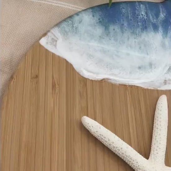 Whale Fin-shaped bamboo charcuterie board with top portion covered in dark and medium blue ocean resin art resembling the waves of the ocean. Laying on a tan beach towel next to a natural starfish (not included) and adorned with orange slices and a matching cheese knife (not included).