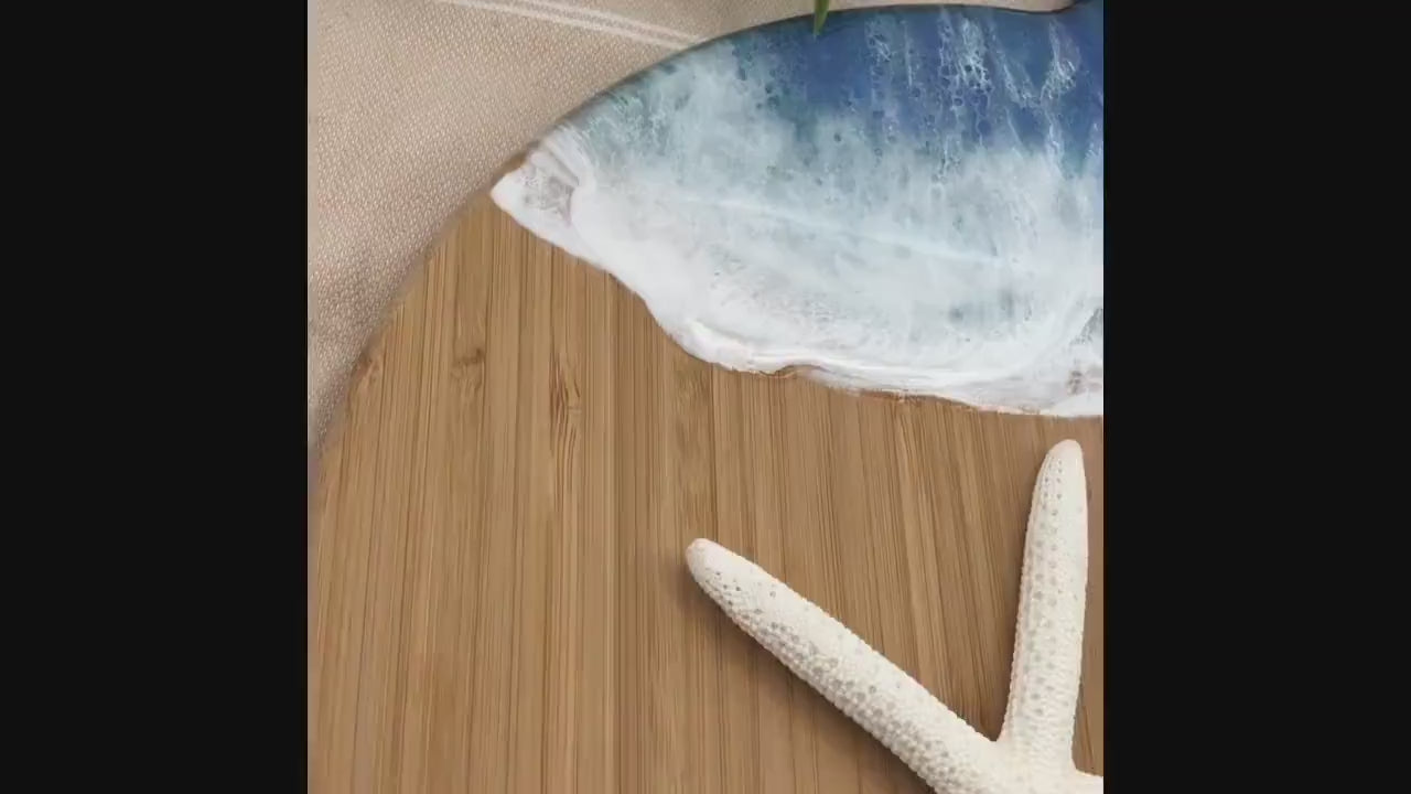 Whale Fin-shaped bamboo charcuterie board with top portion covered in dark and medium blue ocean resin art resembling the waves of the ocean. Laying on a tan beach towel next to a natural starfish (not included) and adorned with orange slices and a matching cheese knife (not included).