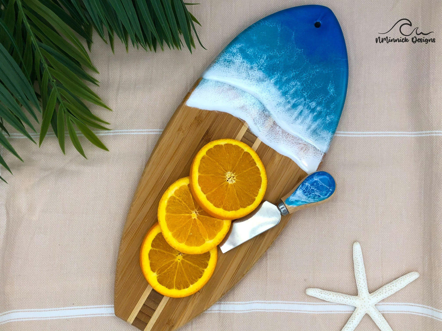 Surfboard-shaped bamboo charcuterie board. Top portion covered in blue and teal ocean resin art resembling the waves of the ocean.  Laying on a tan beach towel next to a natural starfish (not included) and adorned with three orange slices and a matching cheese knife (not included).