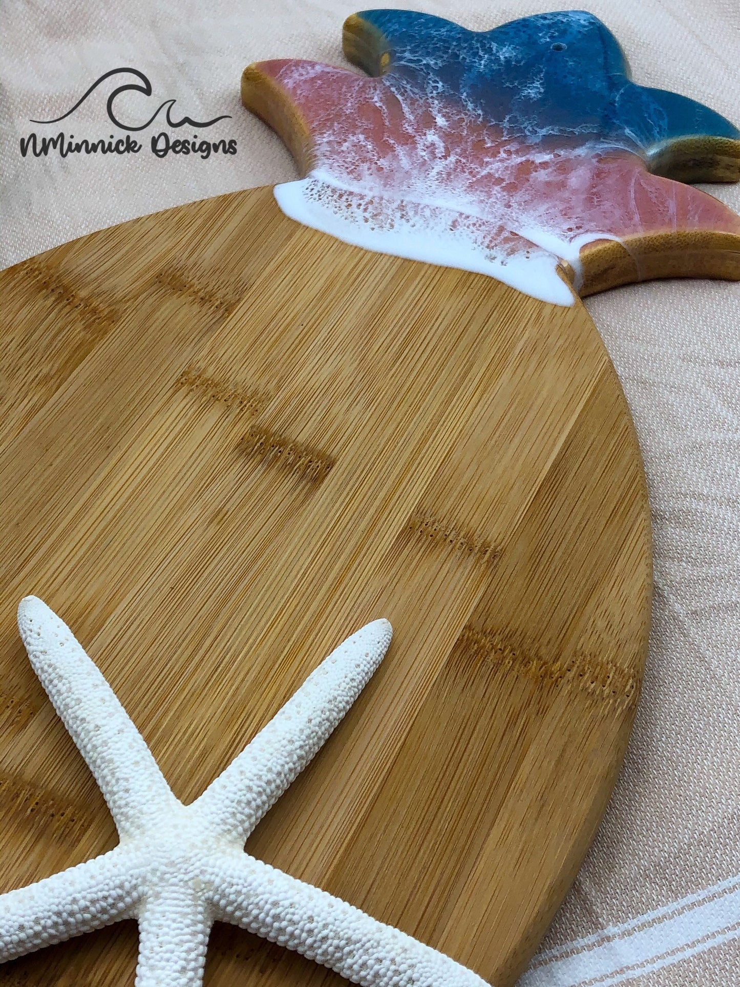 Pineapple-shaped bamboo serving board. Top portion covered in green and pink ocean resin art resembling the waves of the ocean. Laying on a tan beach towel next to a natural starfish (not included).