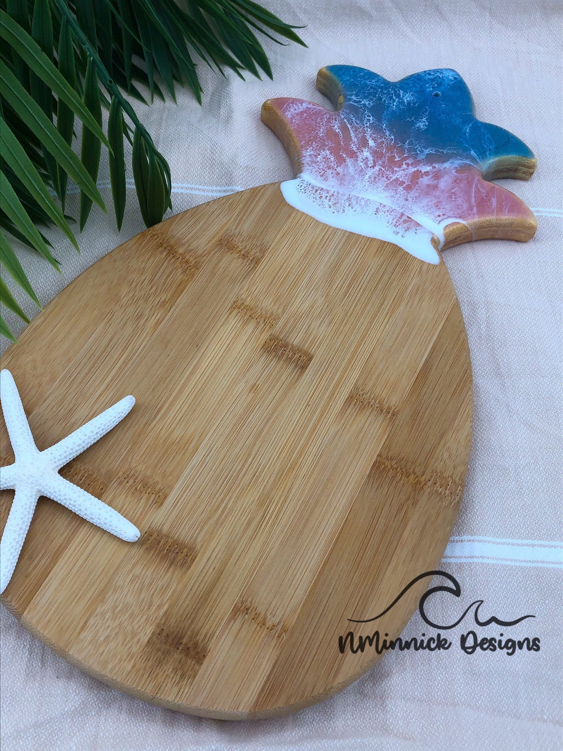Pineapple-shaped bamboo serving board. Top portion covered in green and pink ocean resin art resembling the waves of the ocean. Laying on a tan beach towel next to a natural starfish (not included).