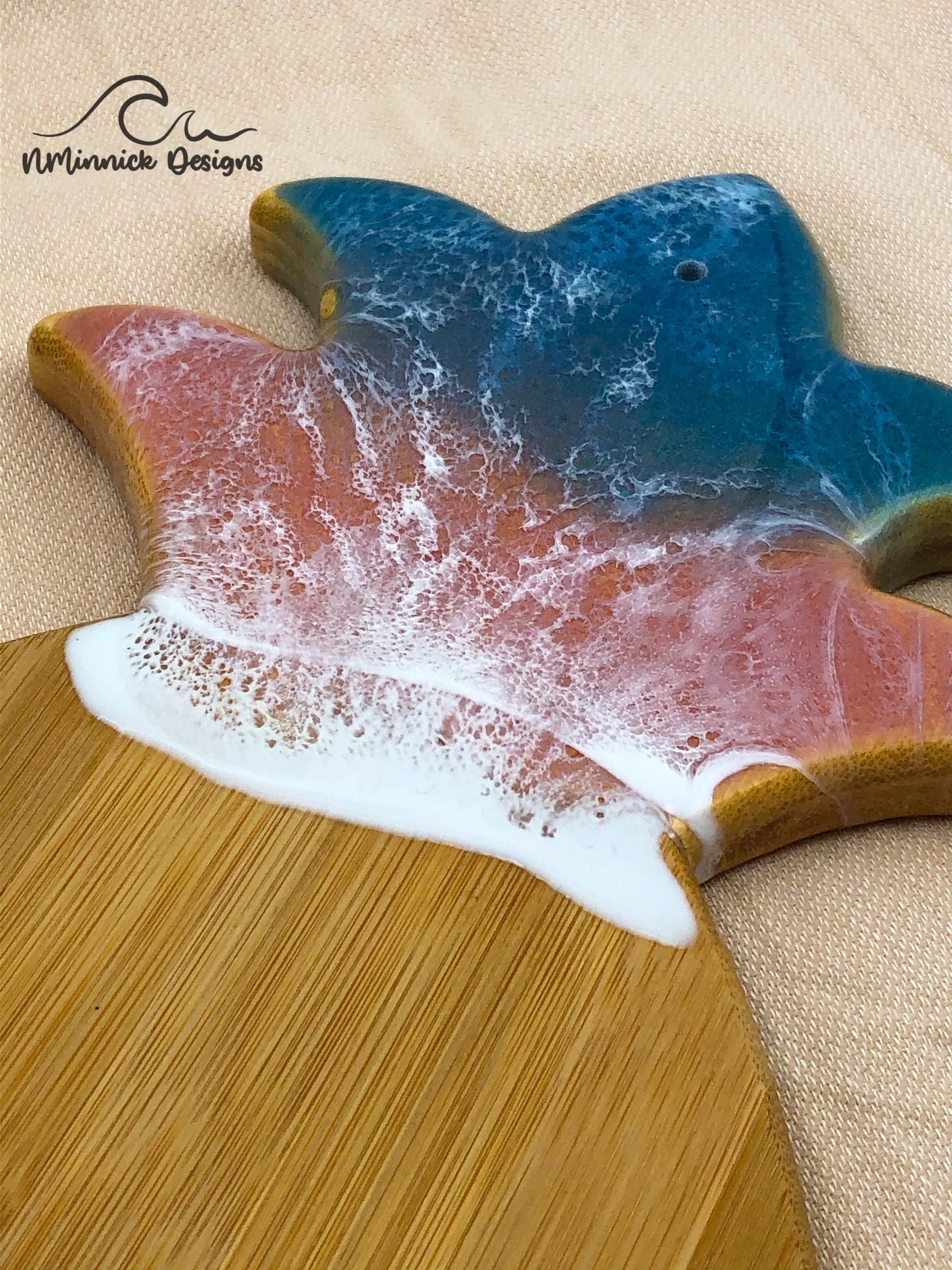 Pineapple-shaped bamboo serving board. Top portion covered in green and pink ocean resin art resembling the waves of the ocean. Laying on a tan beach towel next to a natural starfish (not included).