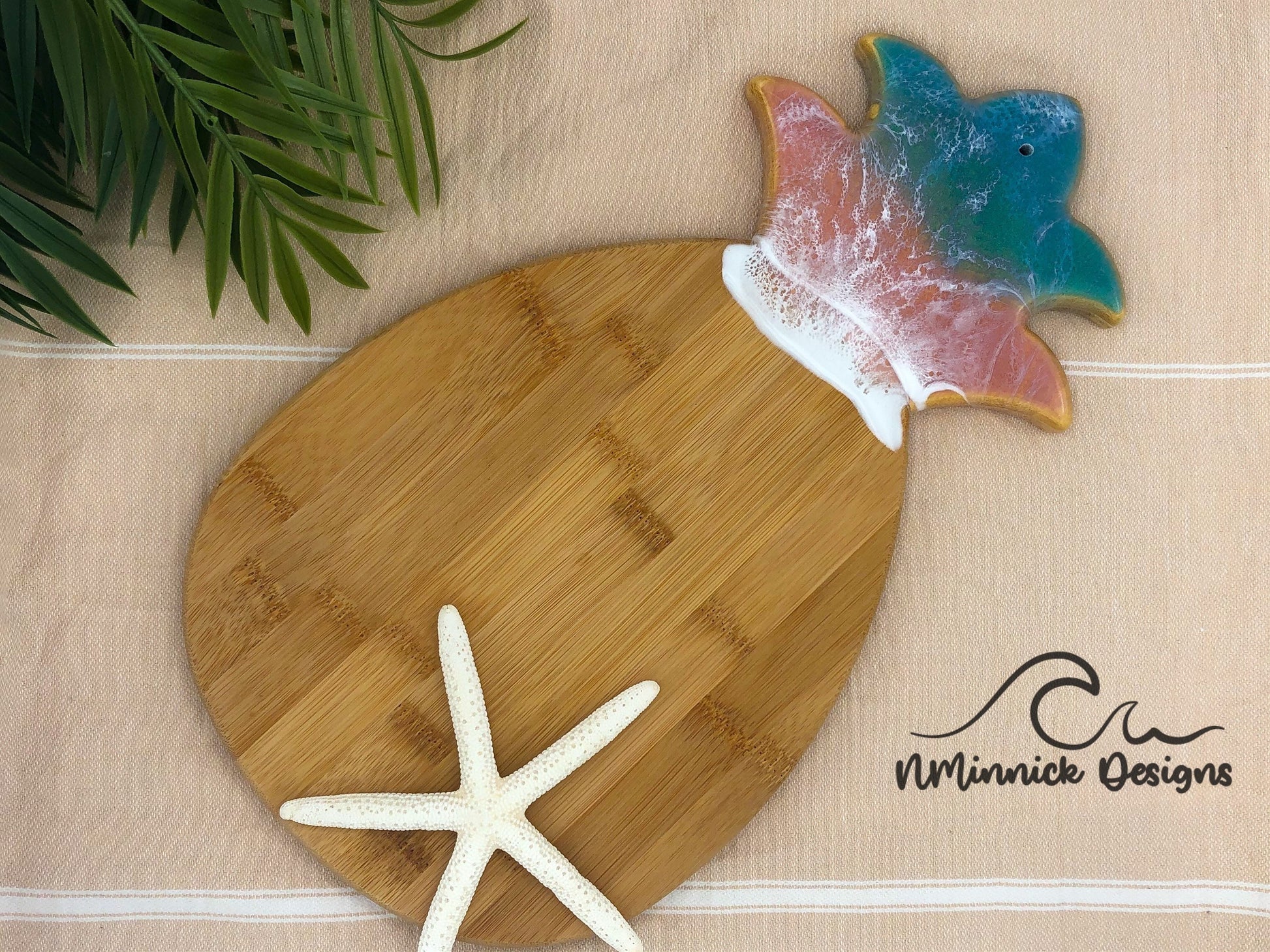 Pineapple-shaped bamboo serving board. Top portion covered in green and pink ocean resin art resembling the waves of the ocean. Laying on a tan beach towel next to a natural starfish (not included).