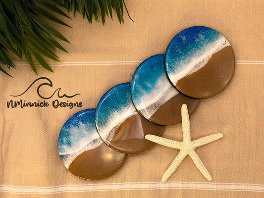 Four, 4-inch round wood coasters covered in a blue and white beach scene made of epoxy resin. Real sand used for the beach on the coasters and cork backings.