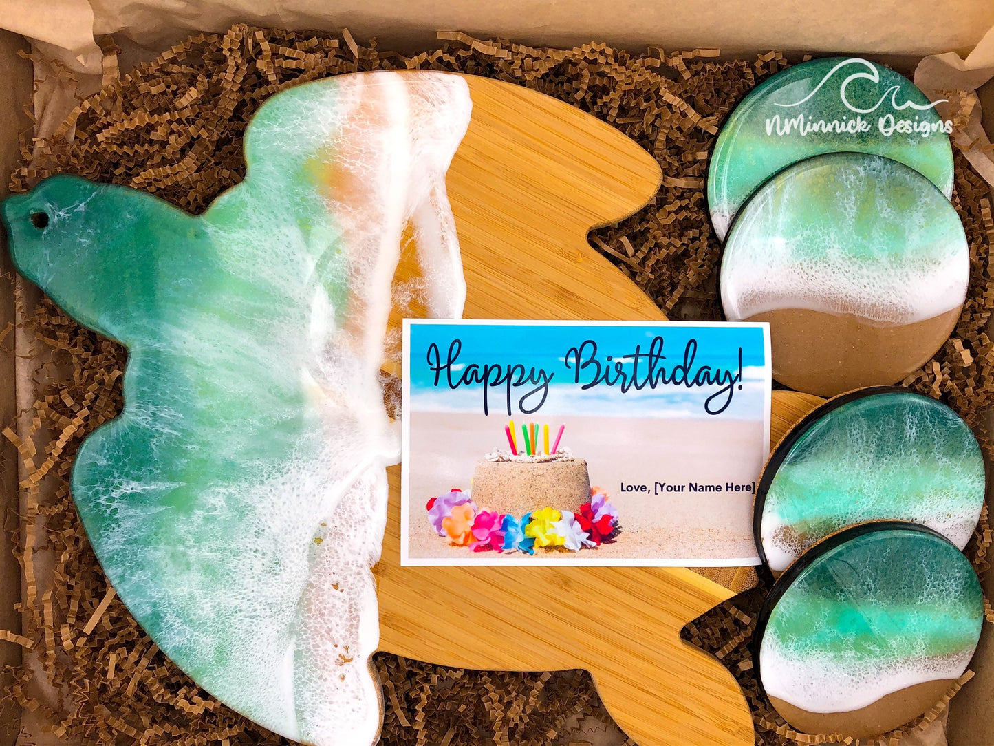 sea turtle-shaped bamboo charcuterie board. Top portion covered in dark green and light green ocean resin art resembling the waves of the ocean.  Matching set of 4 round ocean resin drink coasters with real sand and cork backings.  Shown packaged in a kraft colored eco-friendly and plastic free gift box with a free Happy Birthday card.