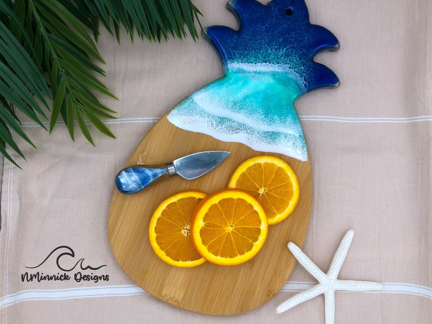 Pineapple-shaped bamboo serving board covered in navy blue and chartreuse ocean resin art to resemble the waves of the ocean.  Laying on a tan beach towel next to a natural starfish.  Orange slices and a matching cheese knife displayed on top.  Cheese knife sold separately. 