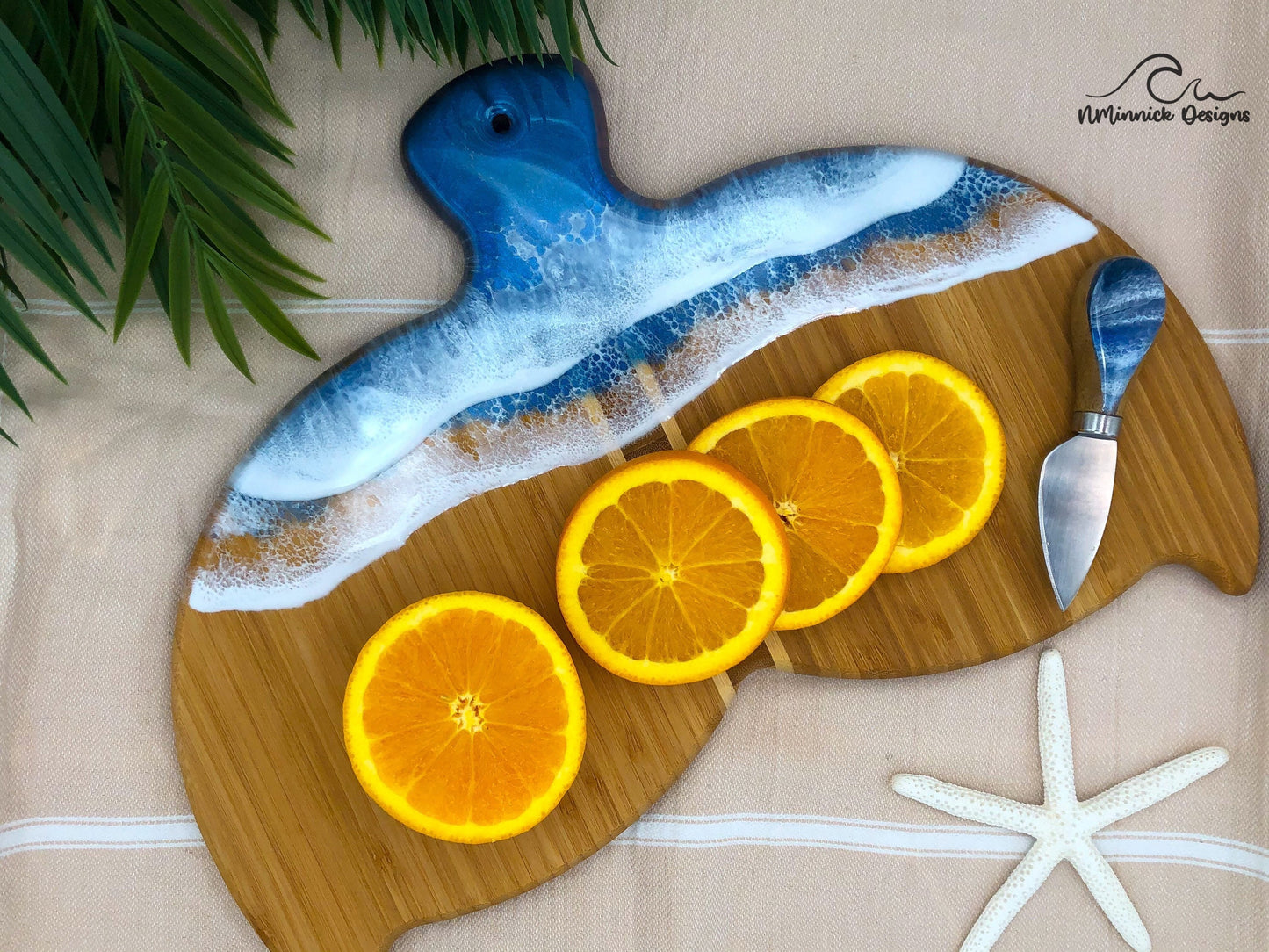 Whale Fin-shaped bamboo charcuterie board with top portion covered in dark and medium blue ocean resin art resembling the waves of the ocean. Laying on a tan beach towel next to a natural starfish (not included) and adorned with orange slices and a matching cheese knife (not included).