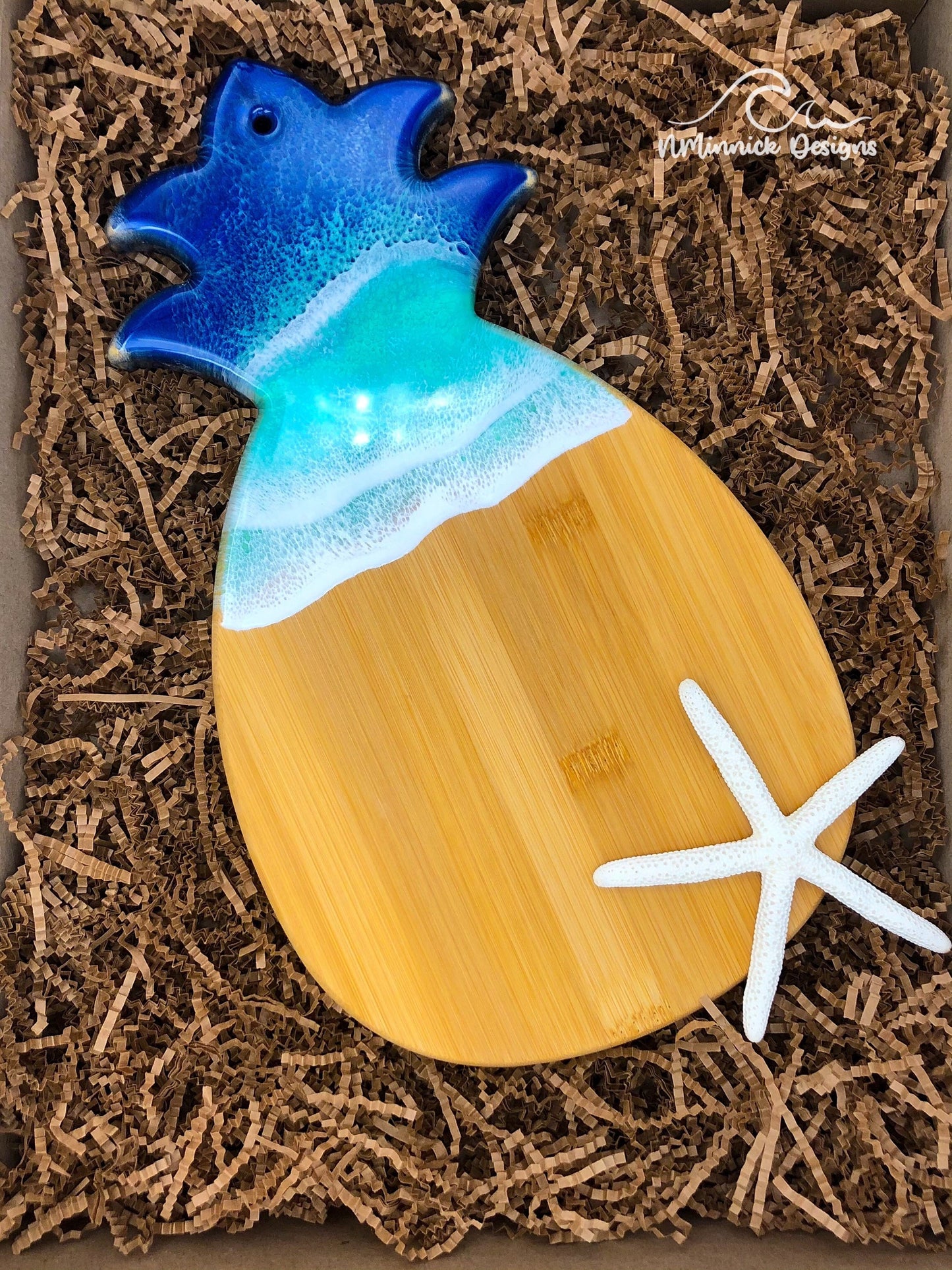 Pineapple shaped bamboo serving board with navy and chartreuse ocean resin art laying in kraft colored eco-friendly and plastic-free packaging next to a natural starfish (not included).