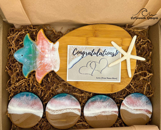 Pineapple-shaped bamboo serving board. Top portion covered in green and pink ocean resin art resembling the waves of the ocean. Matching set of 4 ocean resin drink coasters with real sand and cork backings. Shown packaged in a kraft colored eco-friendly and plastic free gift box with a free Congratulations card.