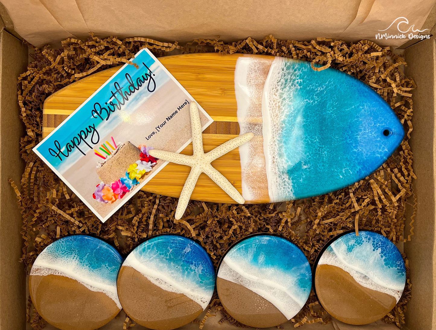 Surfboard-shaped bamboo charcuterie board. Top portion covered in blue and teal ocean resin art resembling the waves of the ocean. Four matching round blue and teal ocean resin drink coasters with real sand and cork backings. Shown packaged in a kraft colored eco-friendly and plastic-free gift box and a free Happy Birthday card.