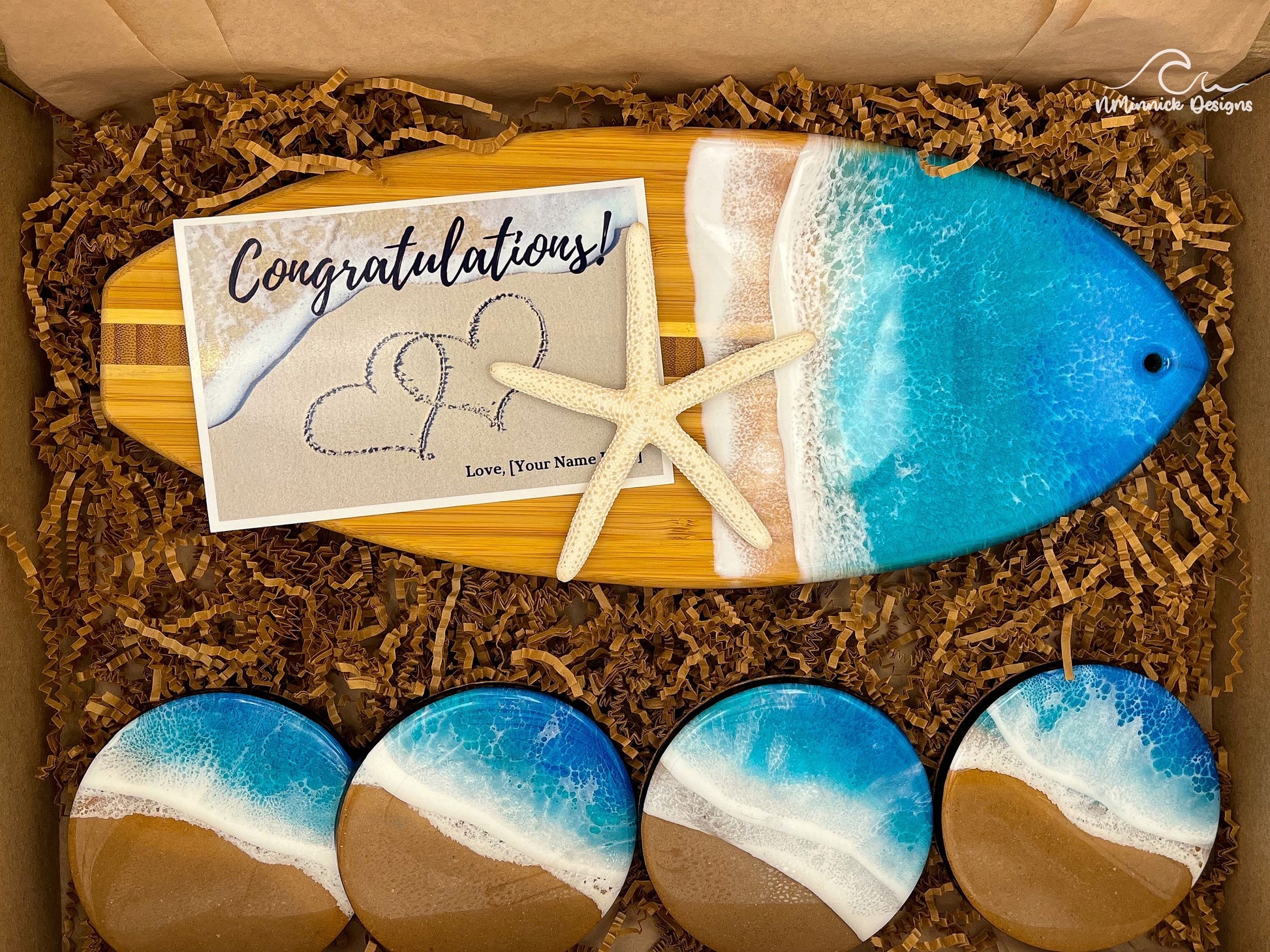 Surfboard-shaped bamboo charcuterie board.  Top portion covered in blue and teal ocean resin art resembling the waves of the ocean.  Four matching round blue and teal ocean resin drink coasters with real sand and cork backings.  Shown packaged in a kraft colored eco-friendly and plastic-free gift box and a free Congratulations card.