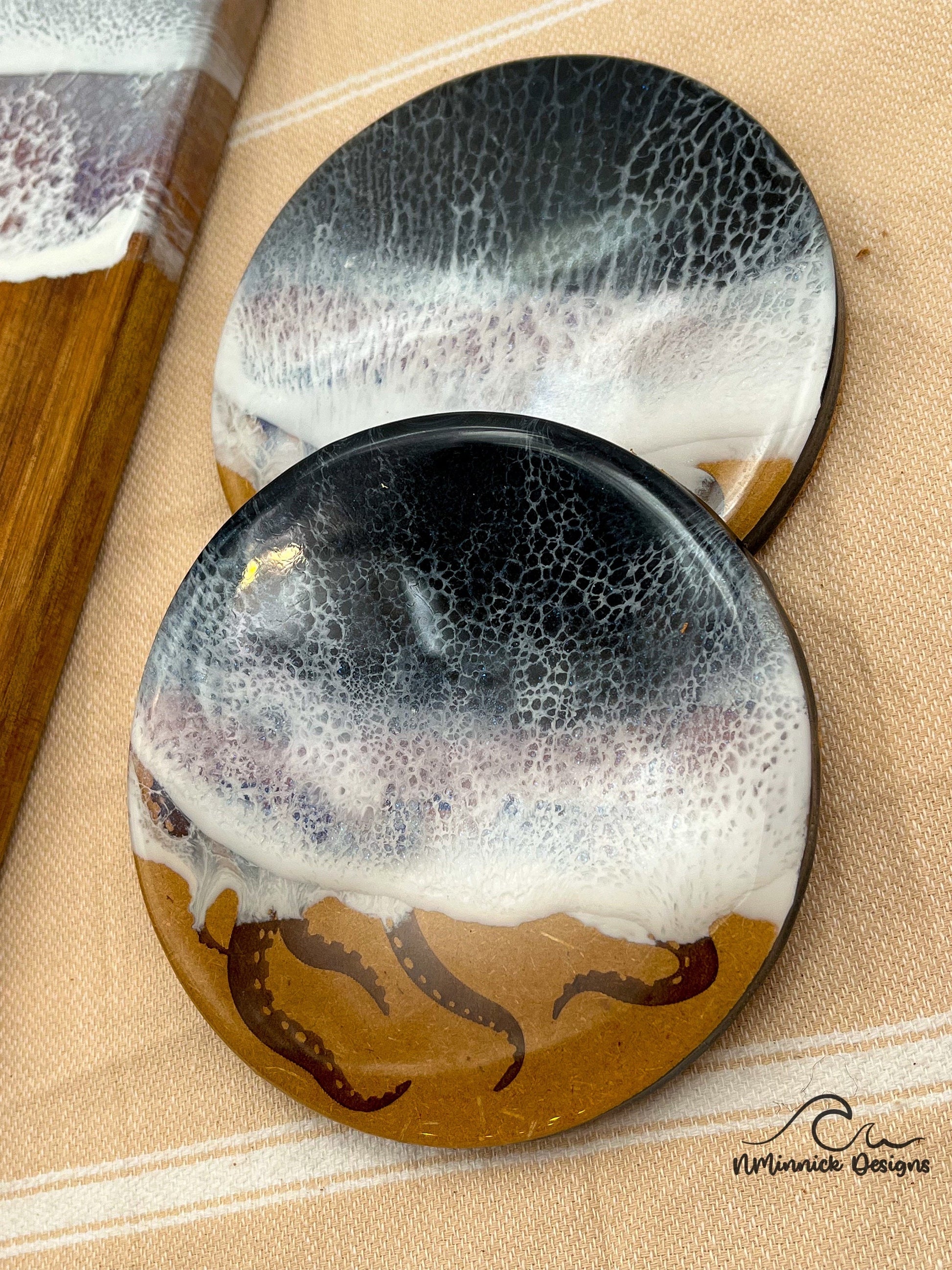 two matching black and silver ocean resin art drink coasters laser engraved with octopus tentacles and coated in epoxy resin with cork backing.