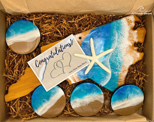 North Carolina-shaped bamboo charcuterie board half covered in blue and teal ocean resin art resembling the waves of the ocean.  Matching set of 4 ocean resin art coasters with cork backings. Free congratulations card. Shown laying in a kraft colored eco-friendly plastic-free gift box.