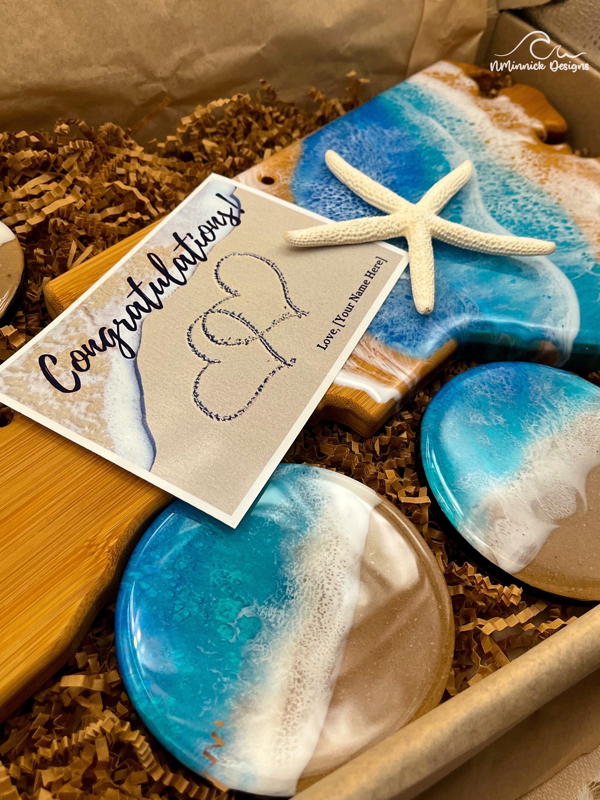 North Carolina-shaped bamboo charcuterie board half covered in blue and teal ocean resin art resembling the waves of the ocean. Matching set of 4 ocean resin art coasters with cork backings. Free congratulations card. Shown laying in a kraft colored eco-friendly plastic-free gift box.